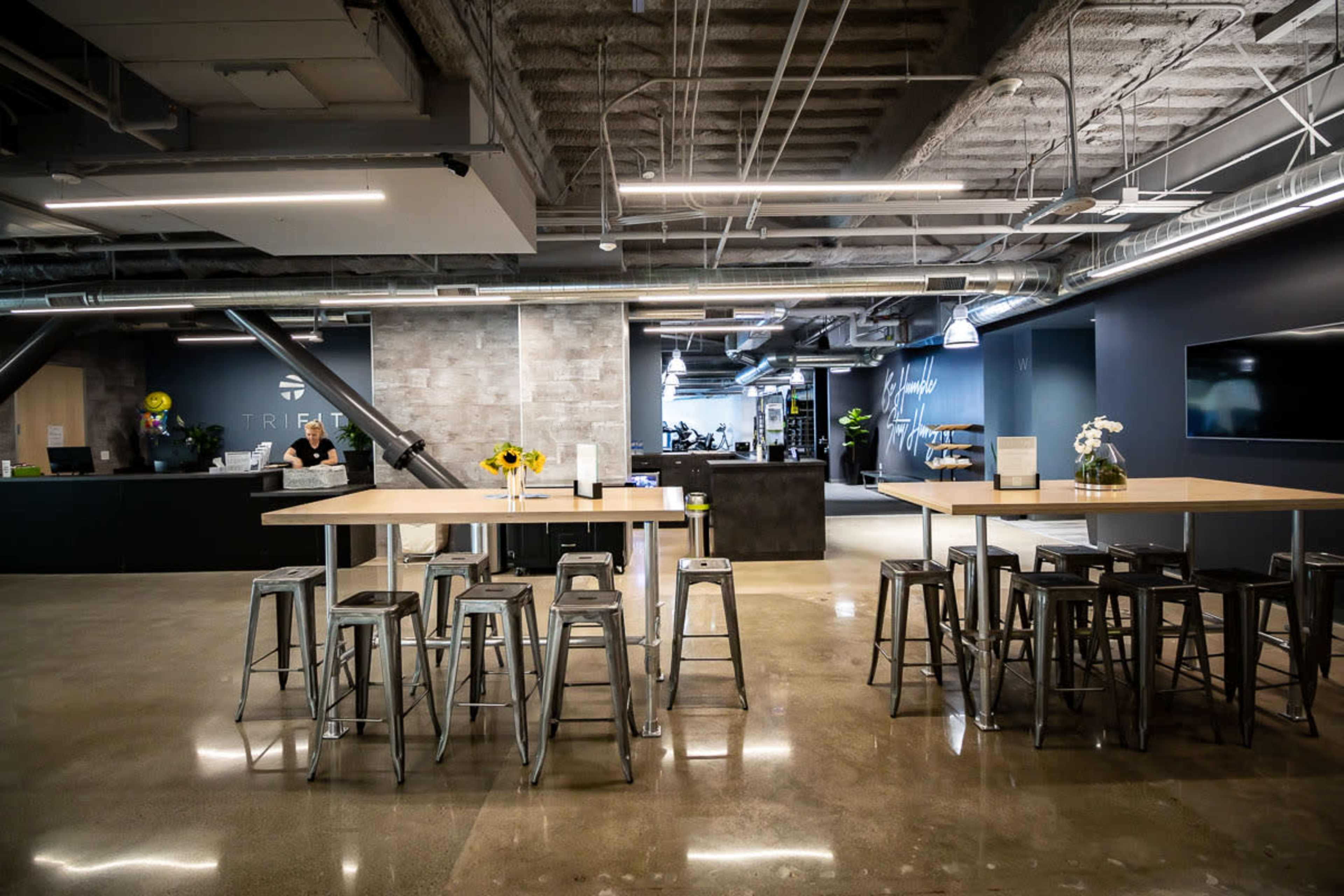 The image shows a modern office space with a large communal table, metal stools, a reception area, and fitness equipment visible in the background.