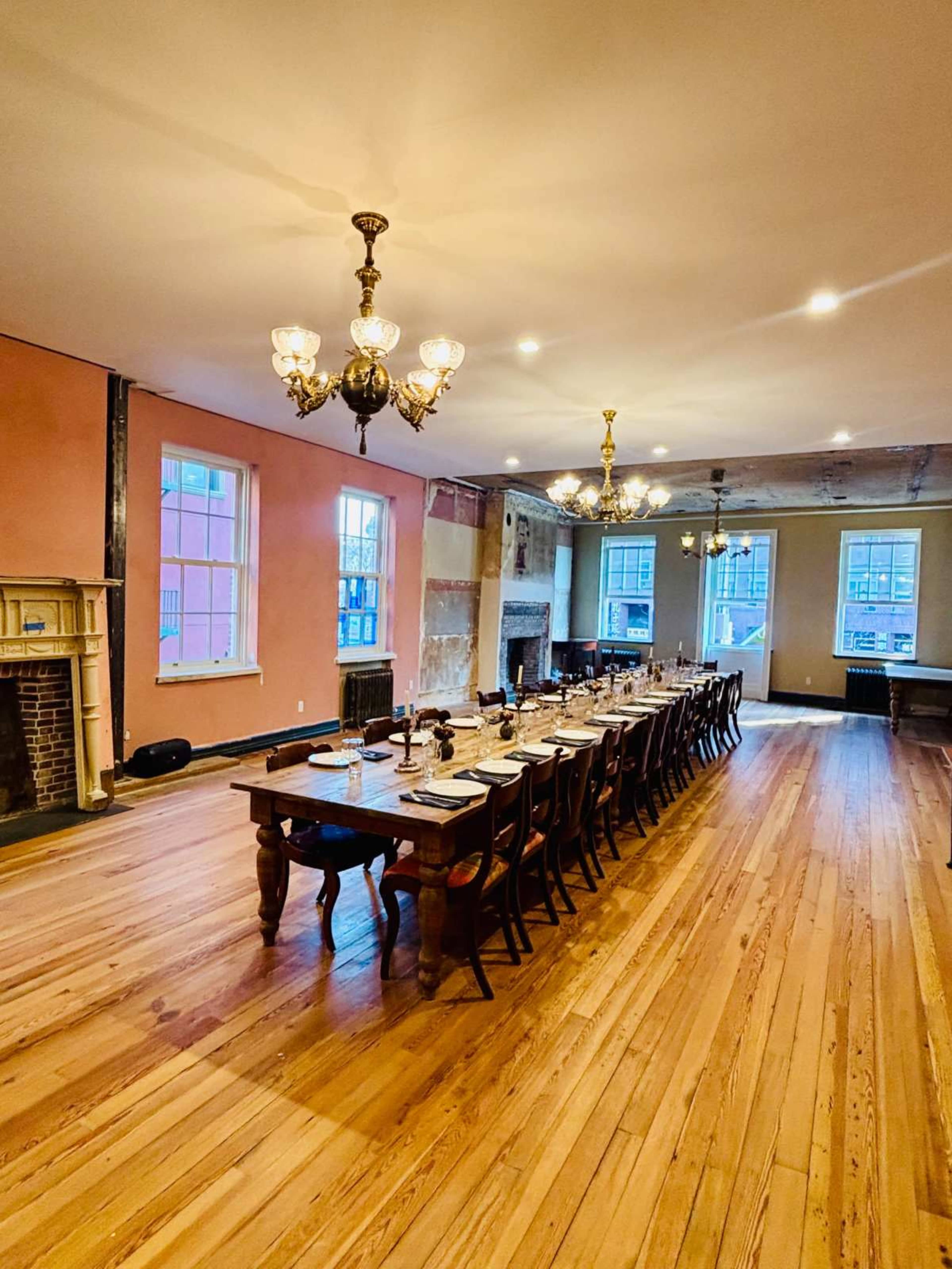 A long wooden dining table is set for a meal in a spacious room with large windows and chandeliers.