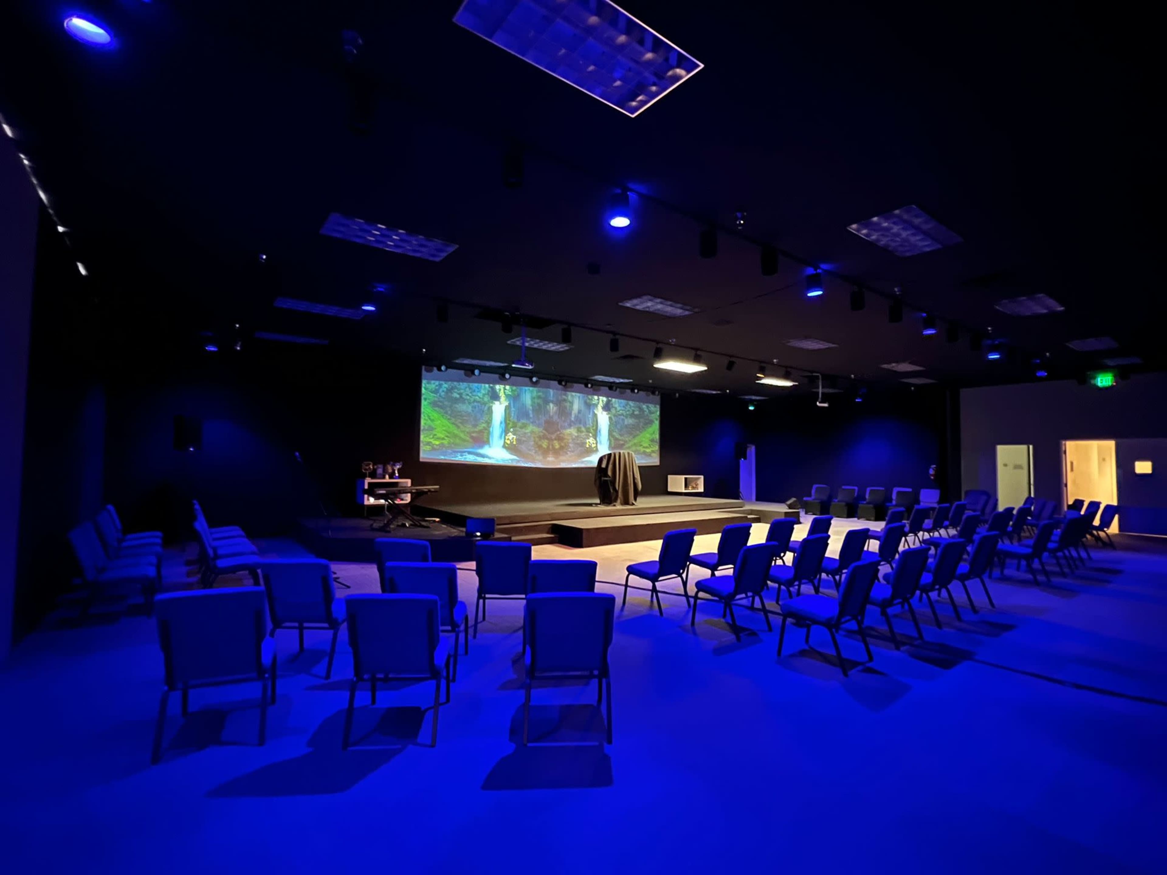 A dark room with blue lighting features rows of empty chairs facing a large projection screen displaying a nature scene.