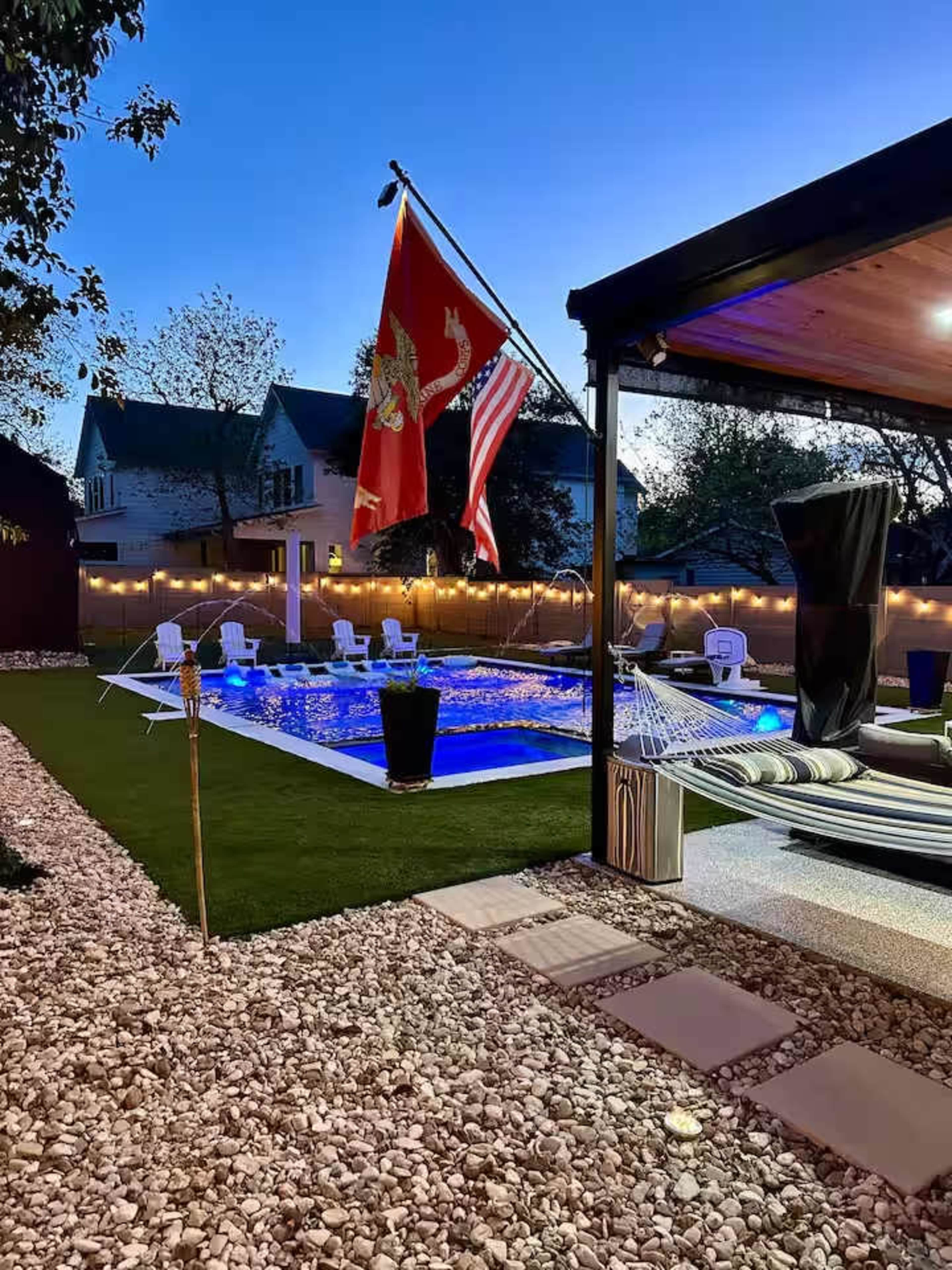 The image shows a well-lit backyard with a swimming pool, surrounded by decorative rocks, lounge chairs, and two flags flying.
