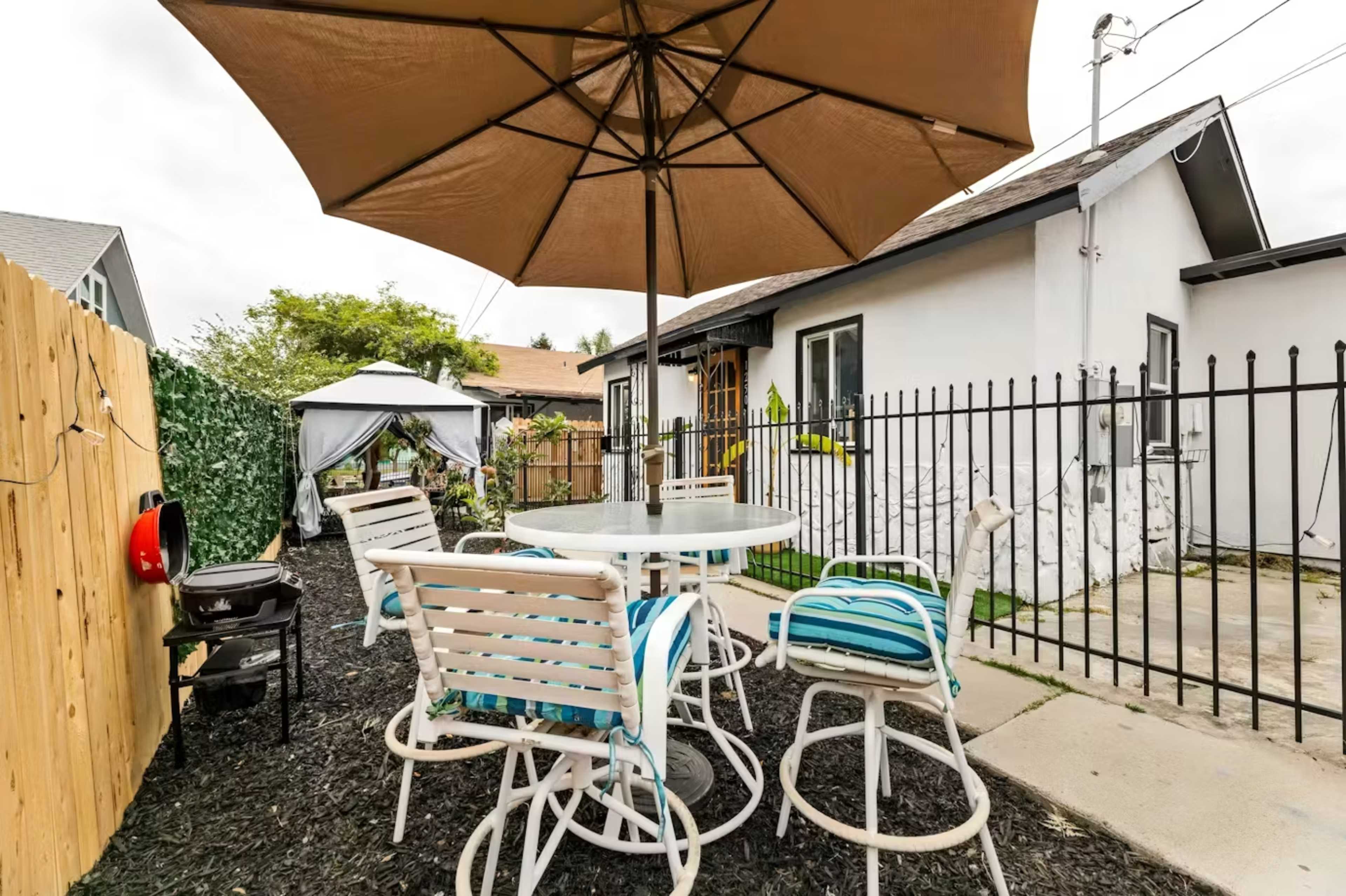 A patio area features a round table with white chairs and a large umbrella, set against a backdrop of a fenced yard and a small house.