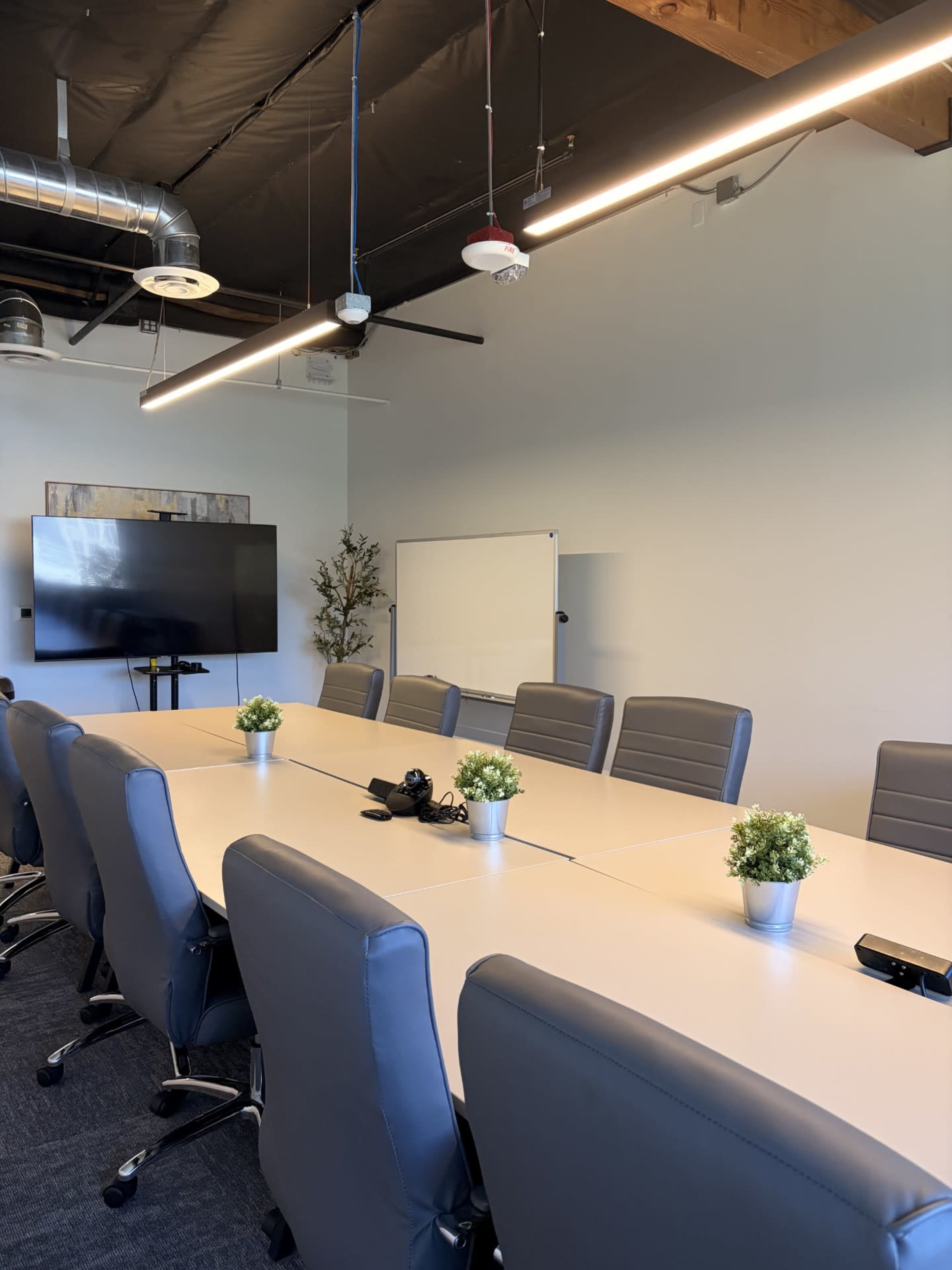 A conference room features a long table surrounded by gray chairs, with a television display and whiteboard on the walls.