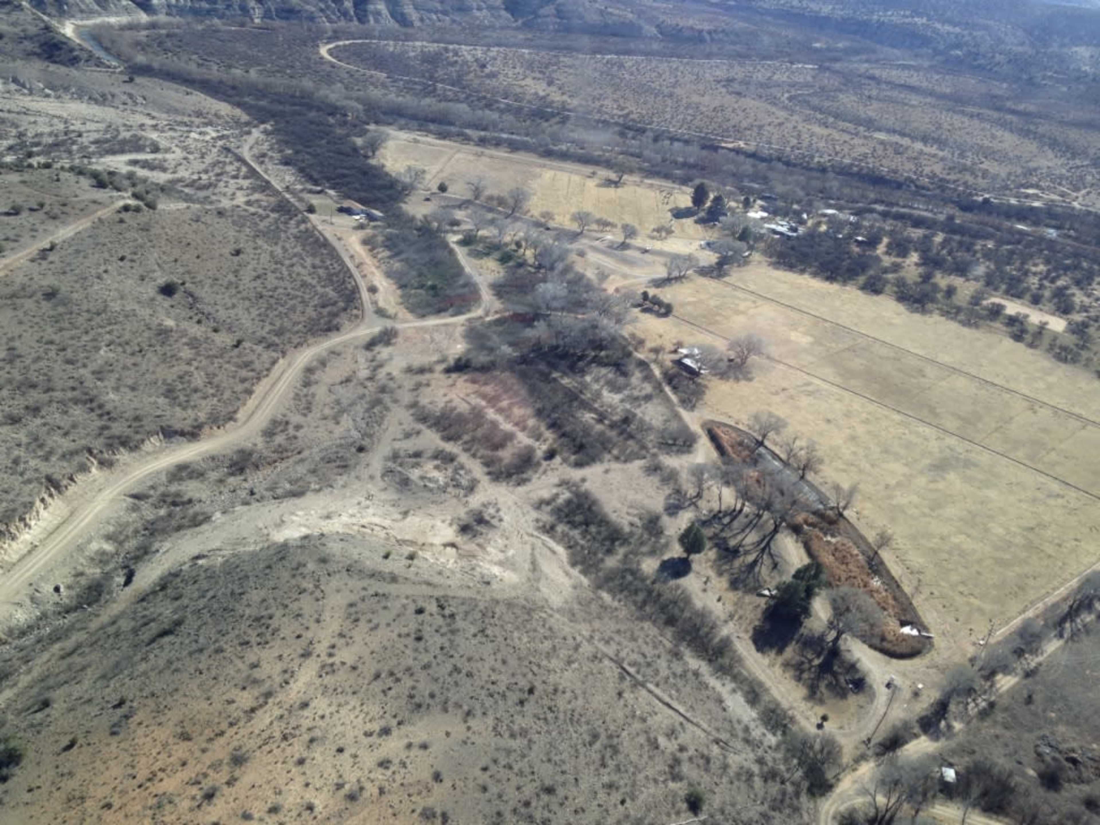 An aerial view shows a landscape with a mix of open fields, scattered trees, a dirt road, and a small cluster of buildings.