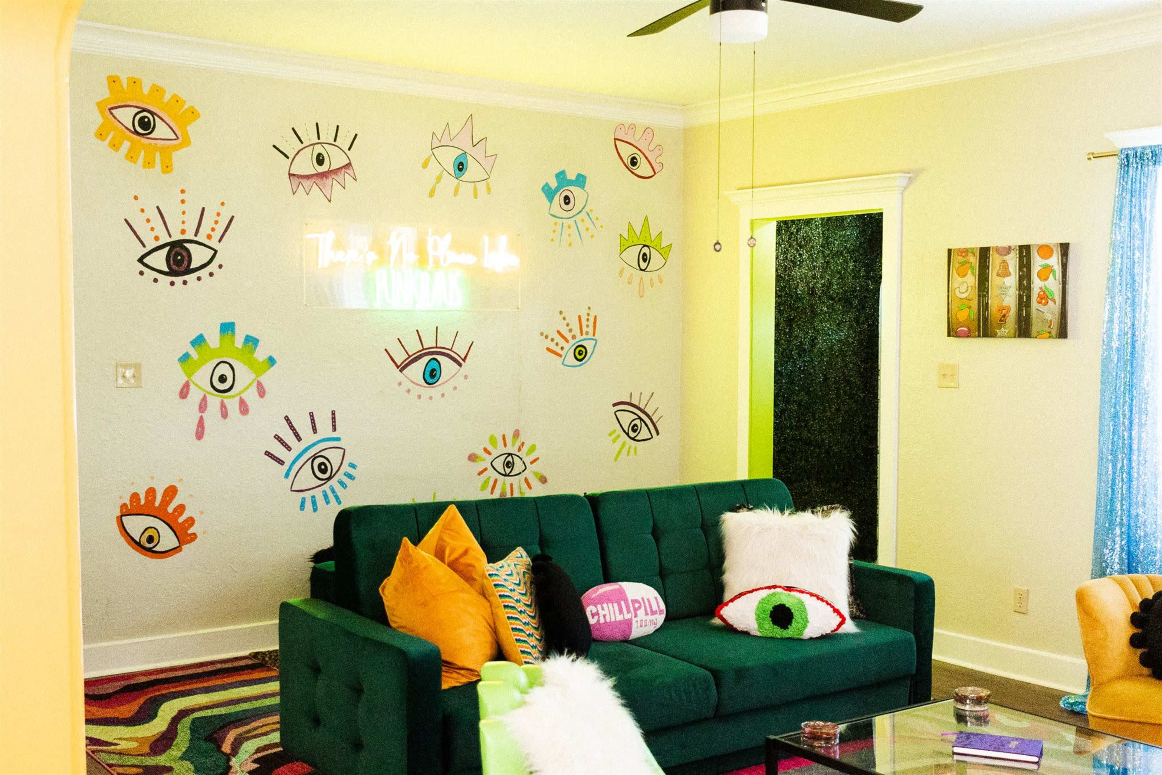 A vibrant living room features a green sofa adorned with decorative pillows, colorful eye-themed wall art, and a neon sign.