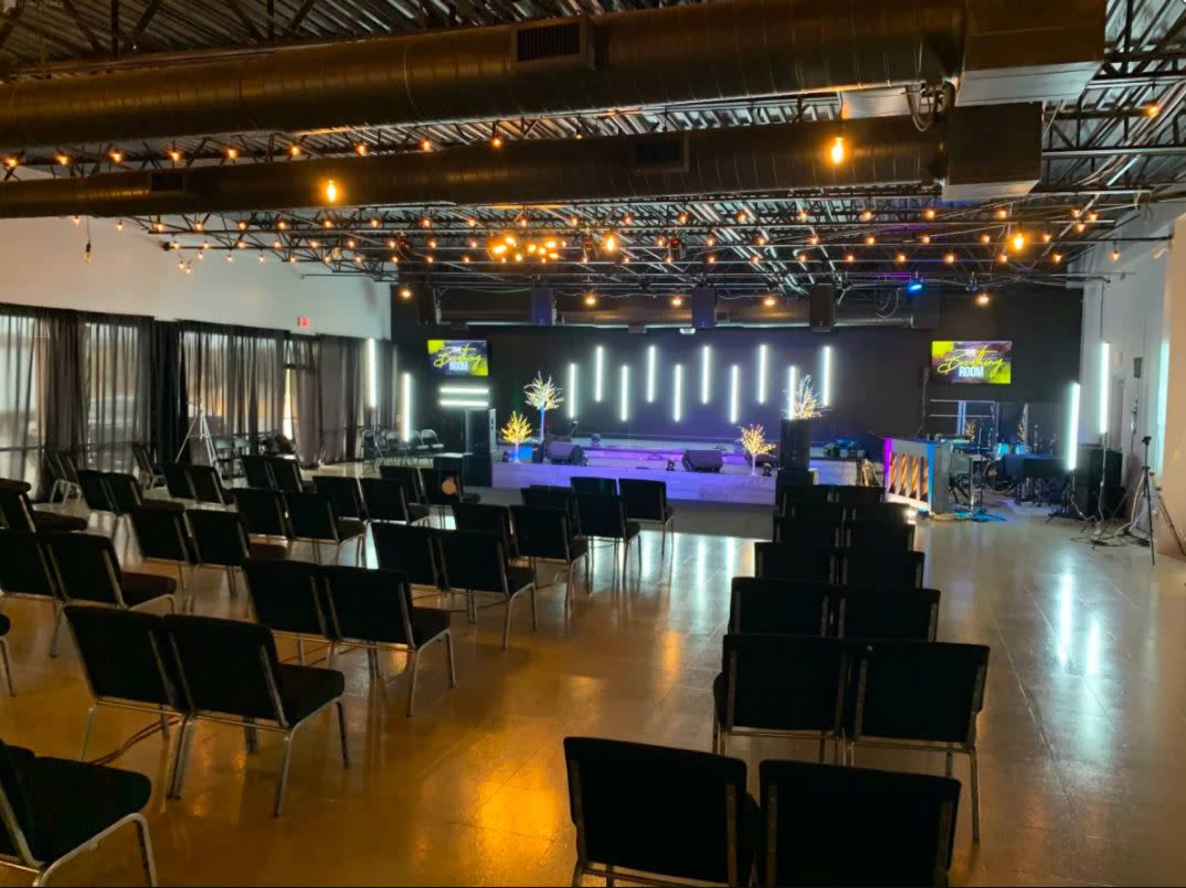 A spacious indoor venue set up for an event, featuring rows of black chairs facing a stage with lighting and audiovisual equipment.