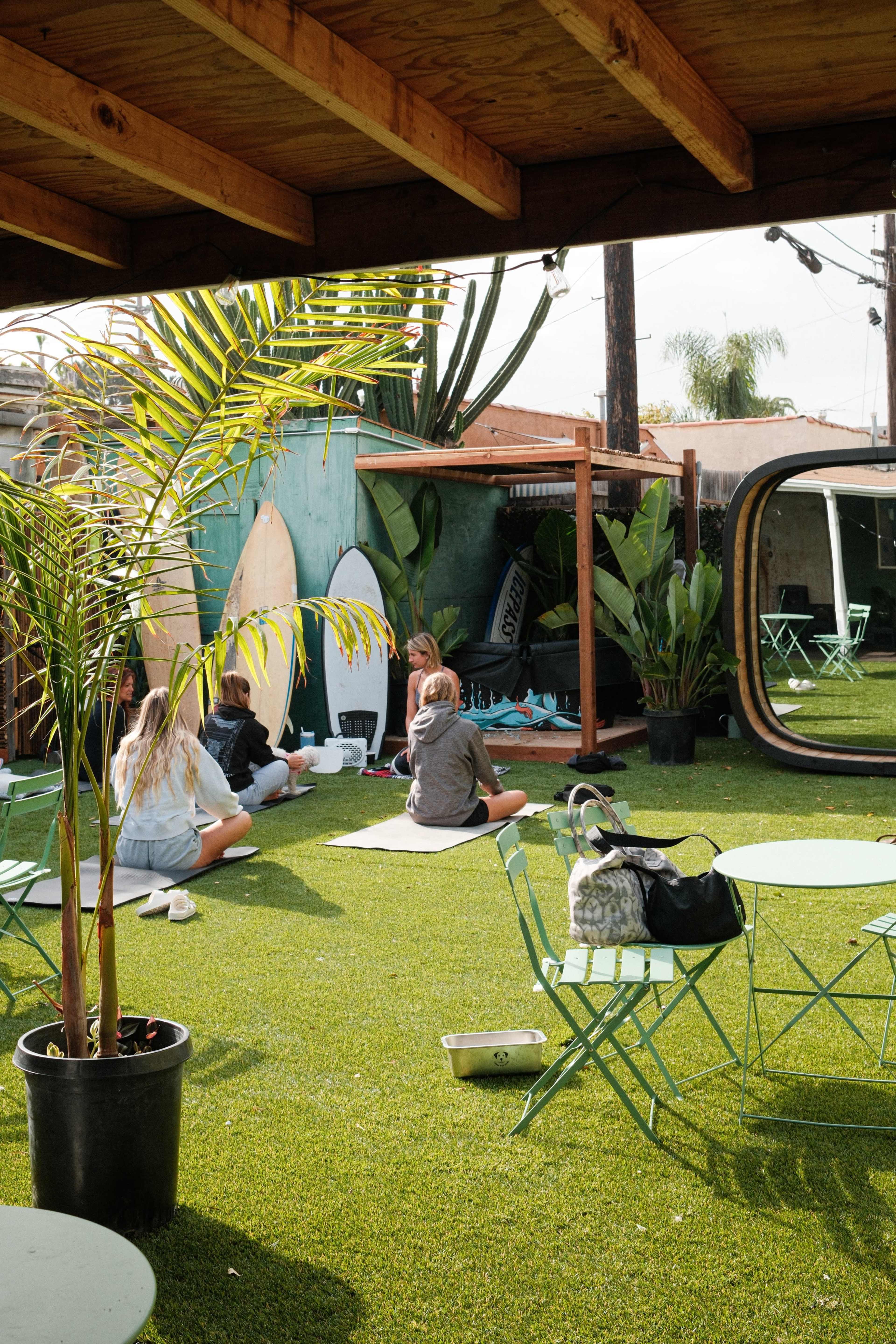 A group of people sits on a grassy area surrounded by surfboards and tropical plants, with green tables and chairs nearby.