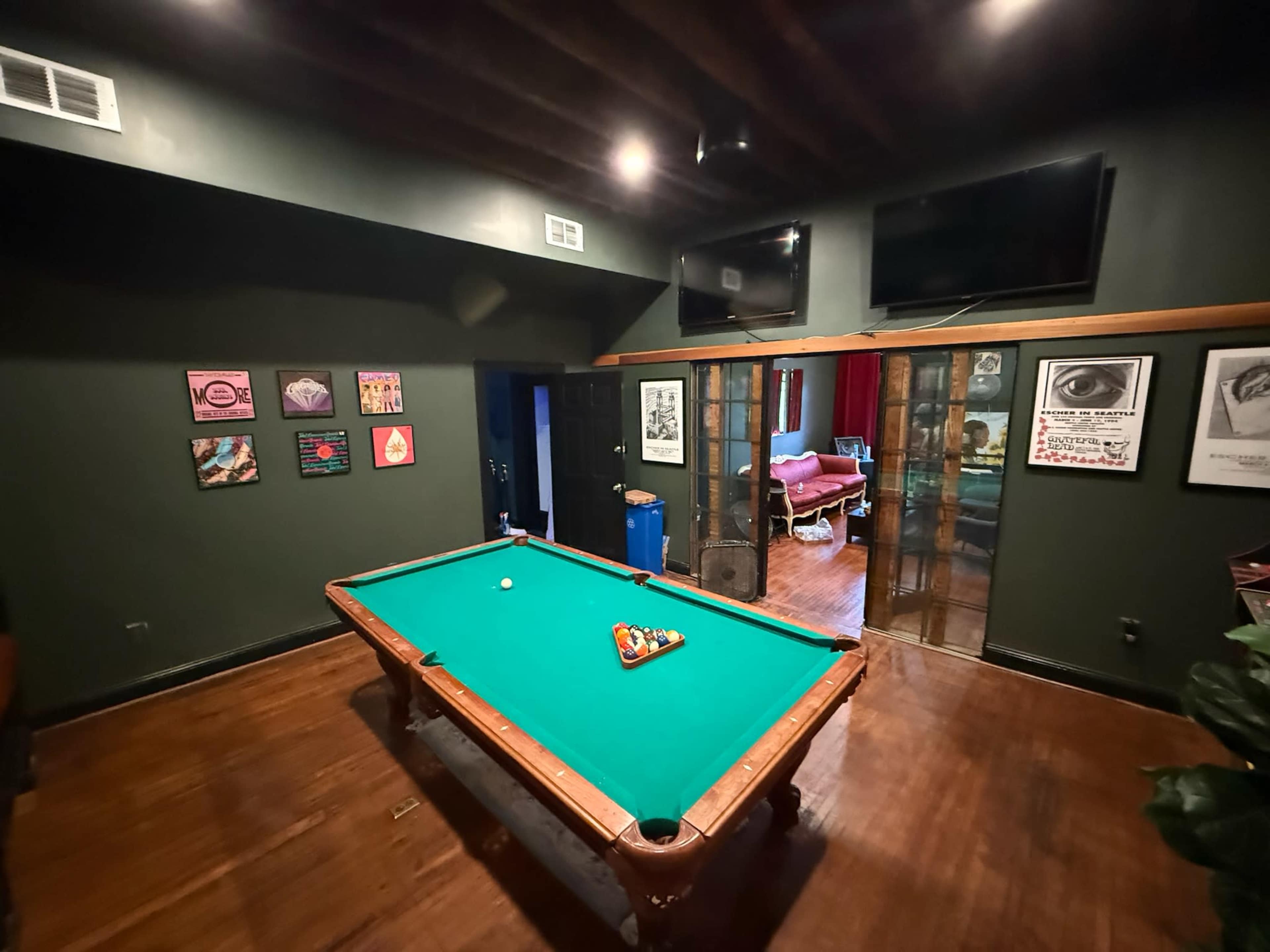 The scene features a green pool table in a dimly lit room with dark green walls, framed artwork on the walls, and two televisions mounted above the table.