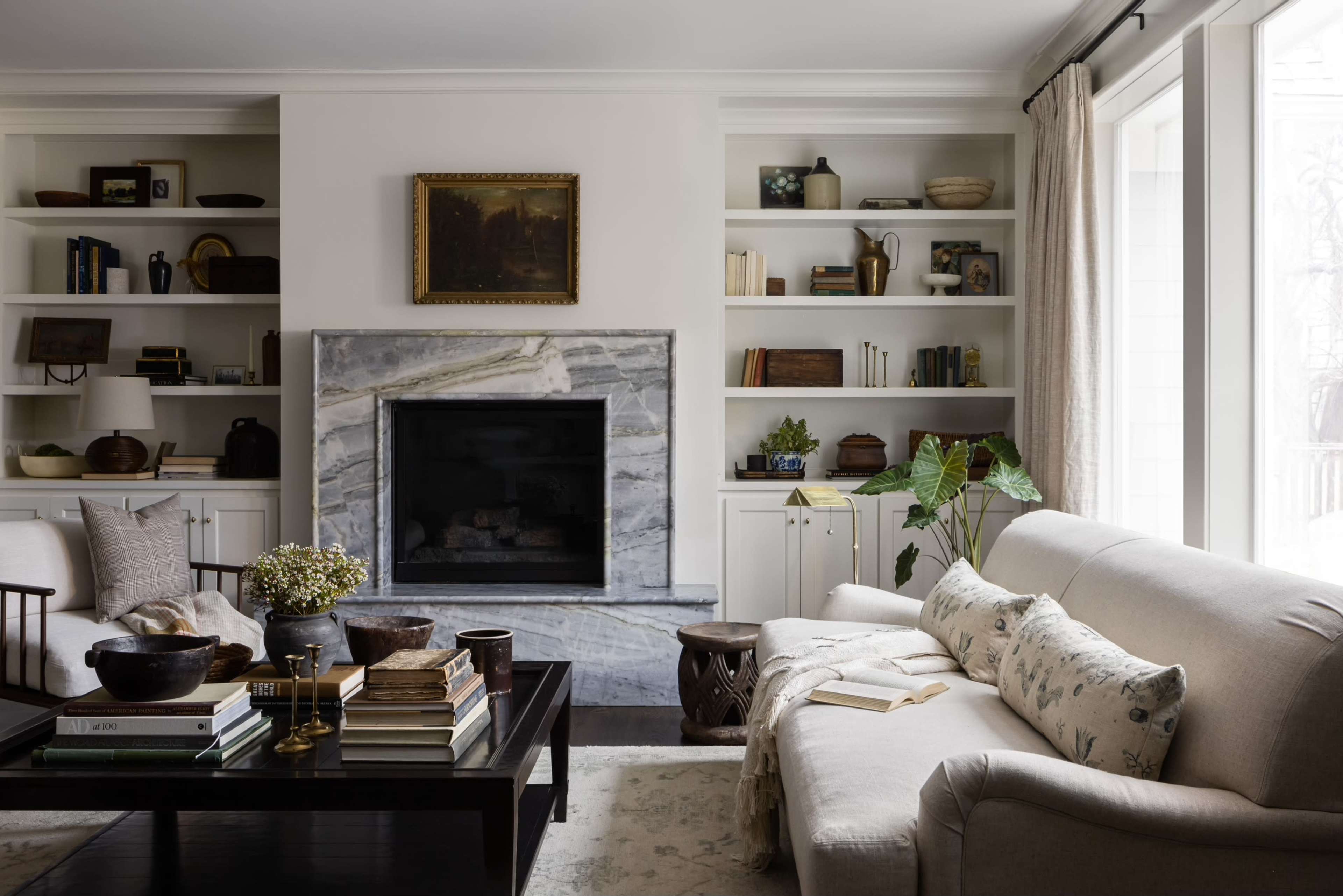 A tidy living room featuring a beige sofa, a marble fireplace, built-in shelves with various books and decor, and large windows allowing natural light.