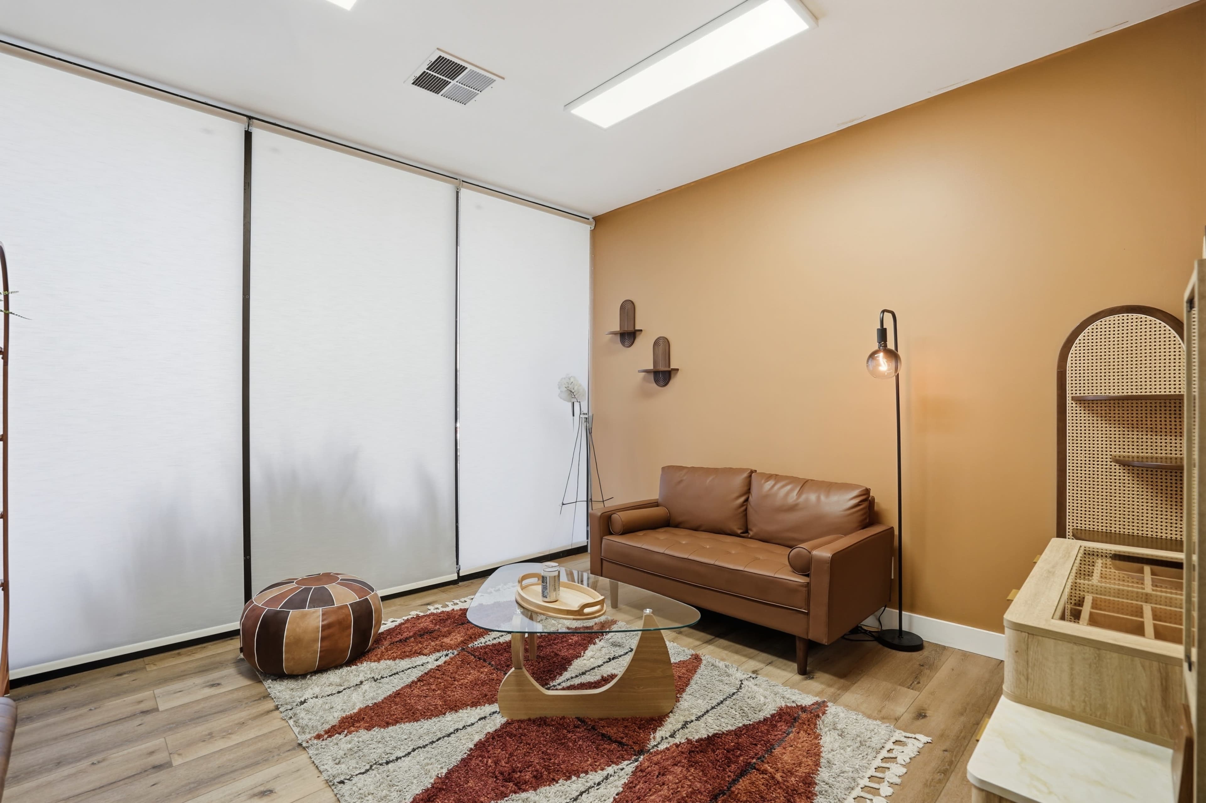 The scene depicts a cozy room with an orange wall, a brown leather sofa, a glass coffee table, and a patterned area rug, complemented by a decorative shelf and a standing fan.