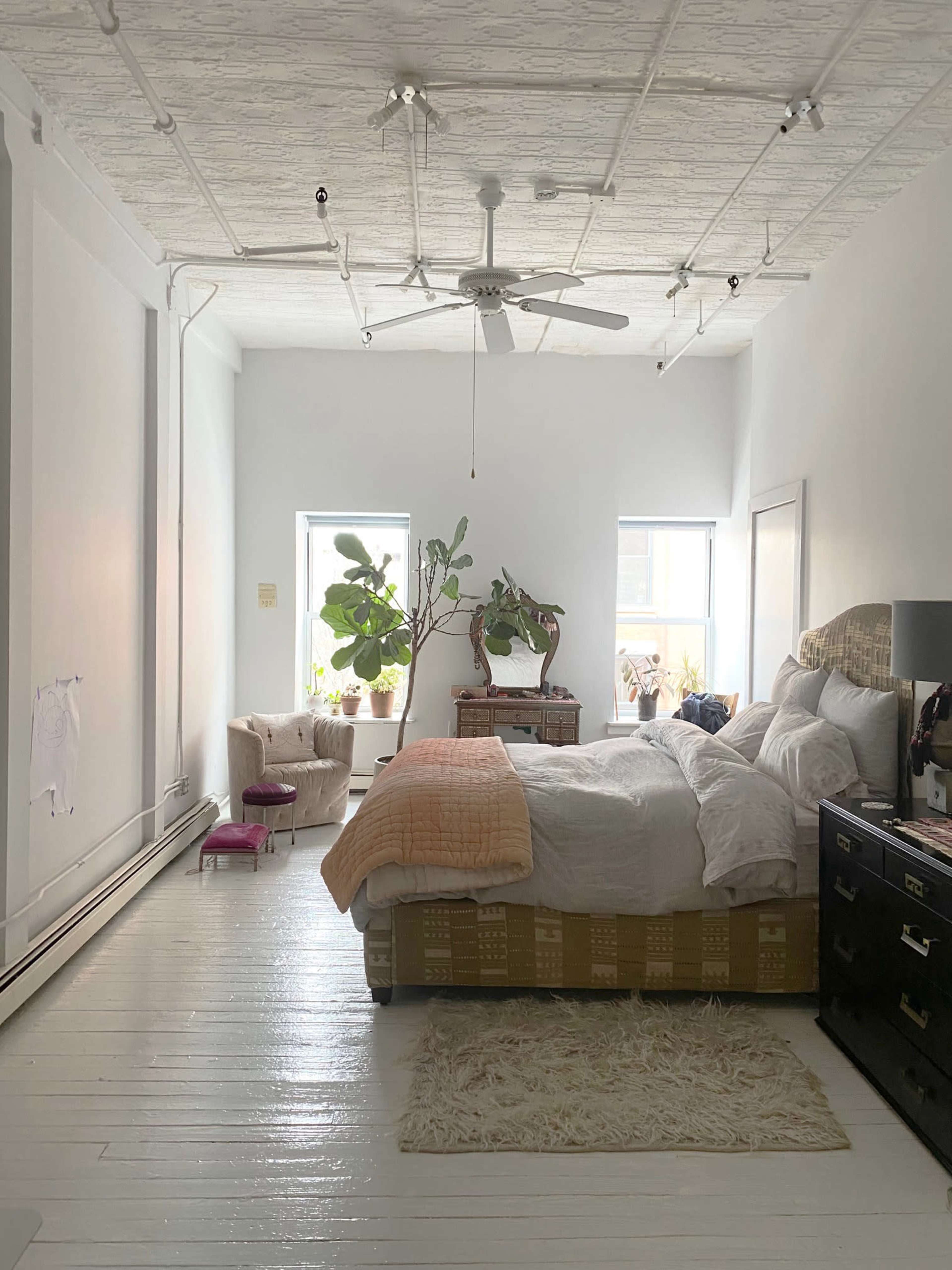 A bright bedroom featuring a large bed with white linens, a dark dresser, two windows, and a couple of potted plants.