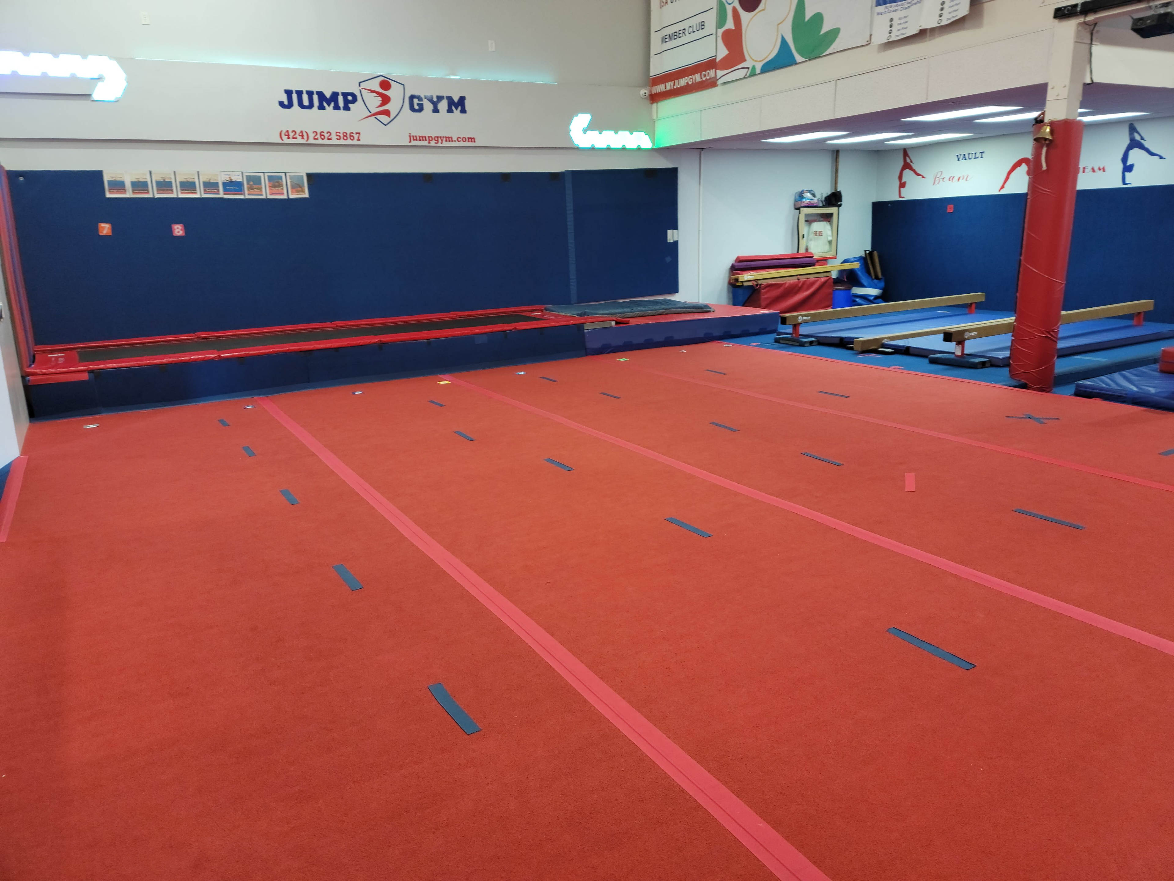 An empty gymnastics gym with red floor mats and blue tape markings on the carpet, along with equipment along the walls.