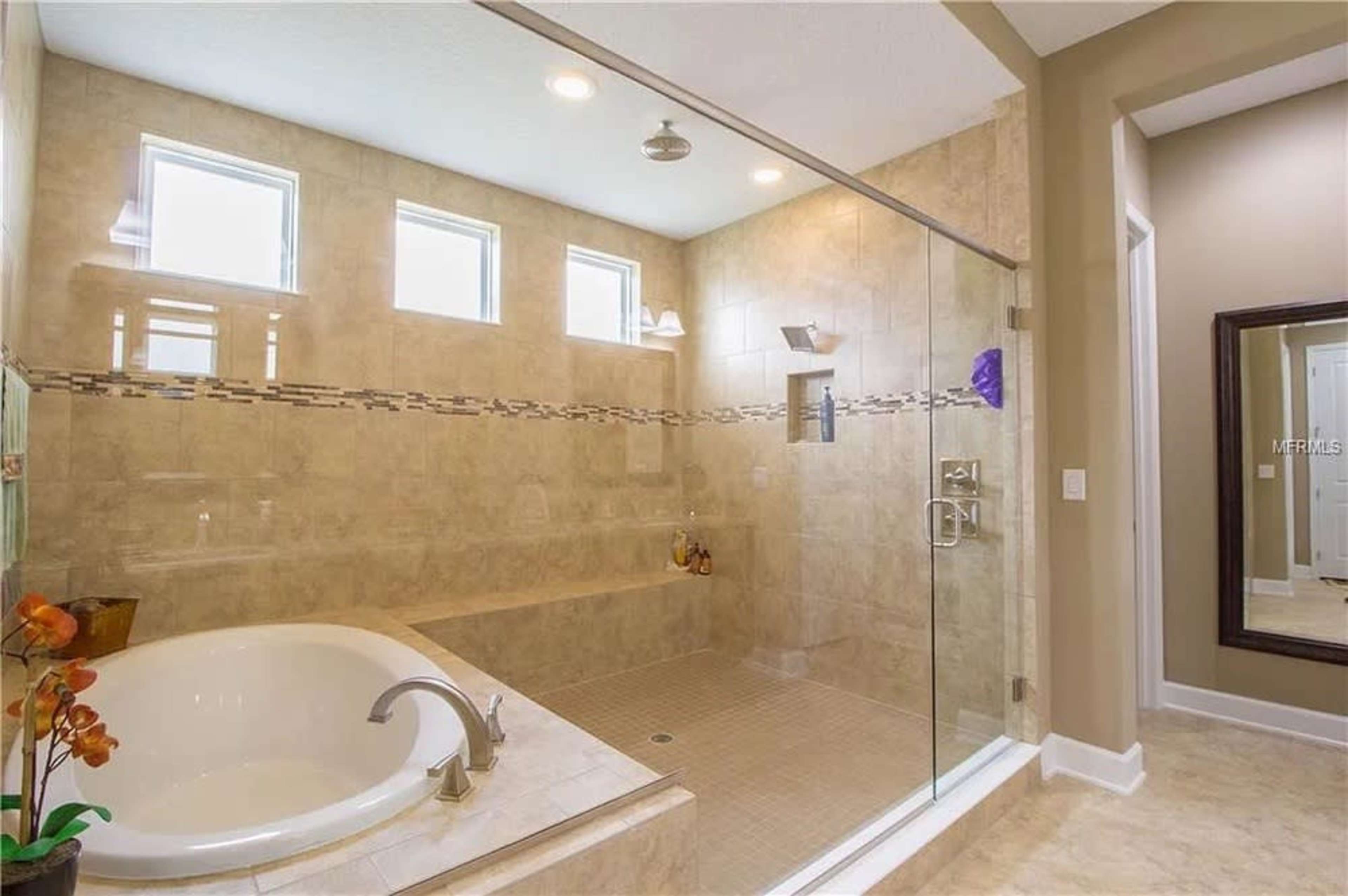 The image shows a spacious bathroom featuring a glass-enclosed shower, a white soaking tub, and natural light coming through multiple windows.