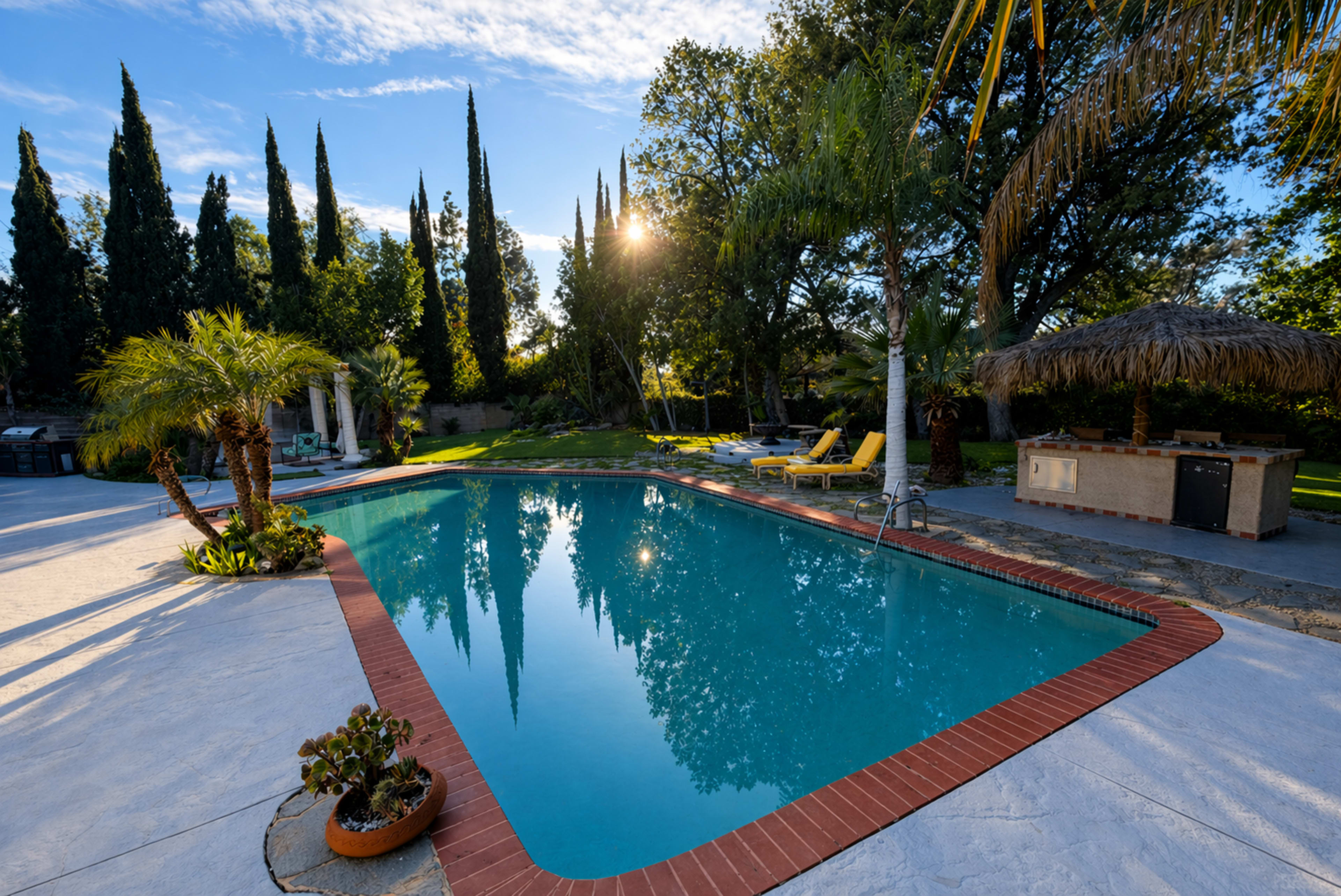 The image shows a clear swimming pool surrounded by palm trees, lounge chairs, and a cabana, with sunlight filtering through the trees in the background.
