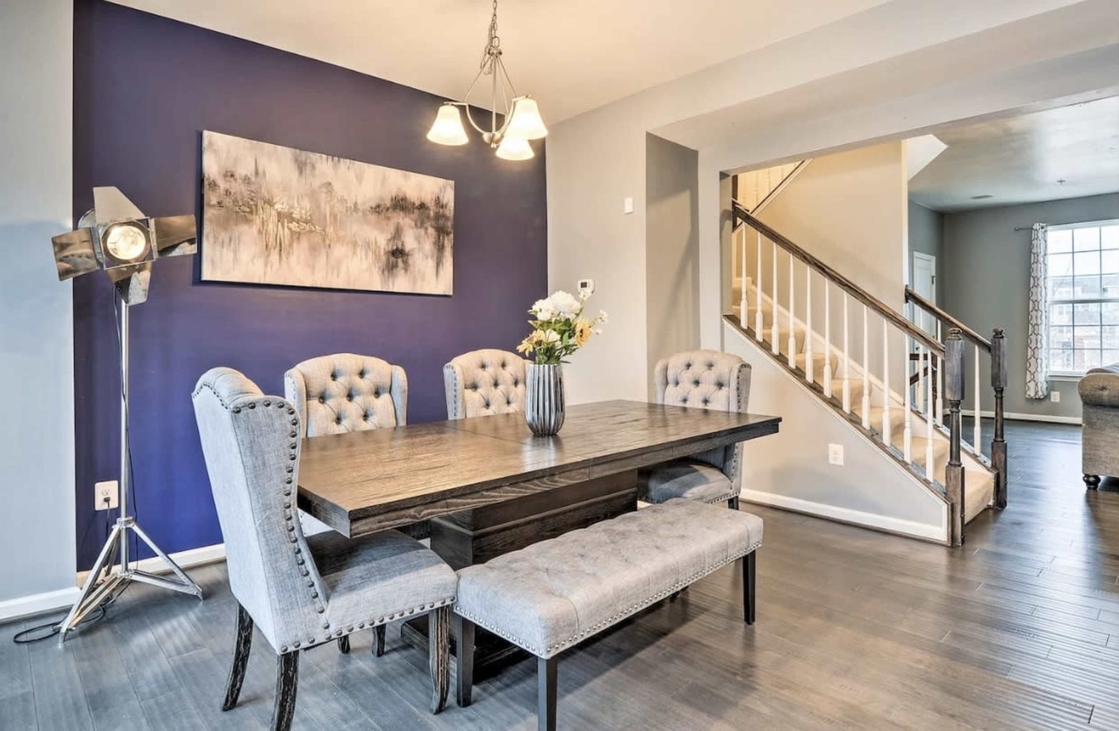 A dining area featuring a wooden table surrounded by upholstered chairs and a bench, with a wall art piece and a staircase in the background.