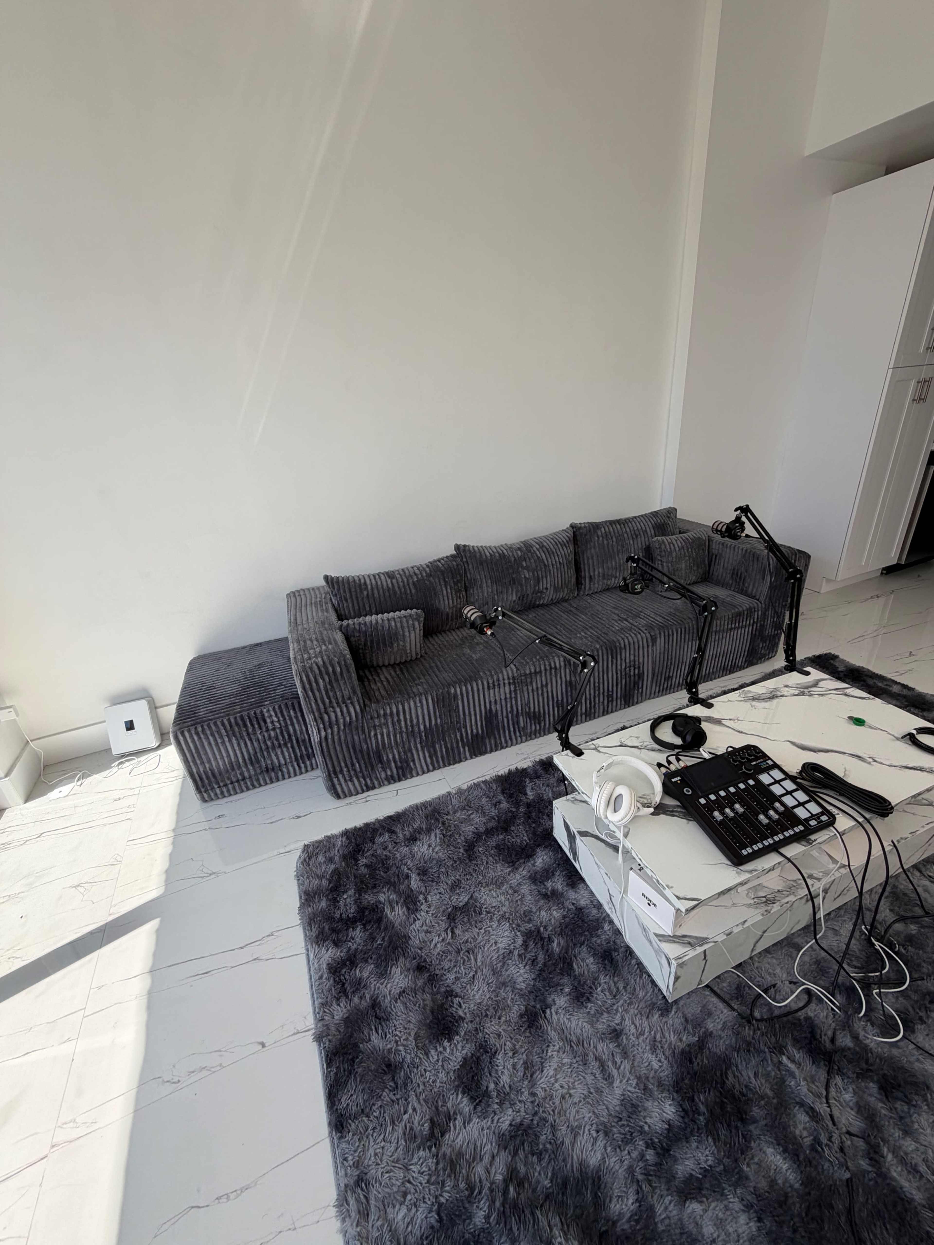 The image shows a modern minimalistic room with a gray striped sofa, a large gray rug, and recording equipment on a table.