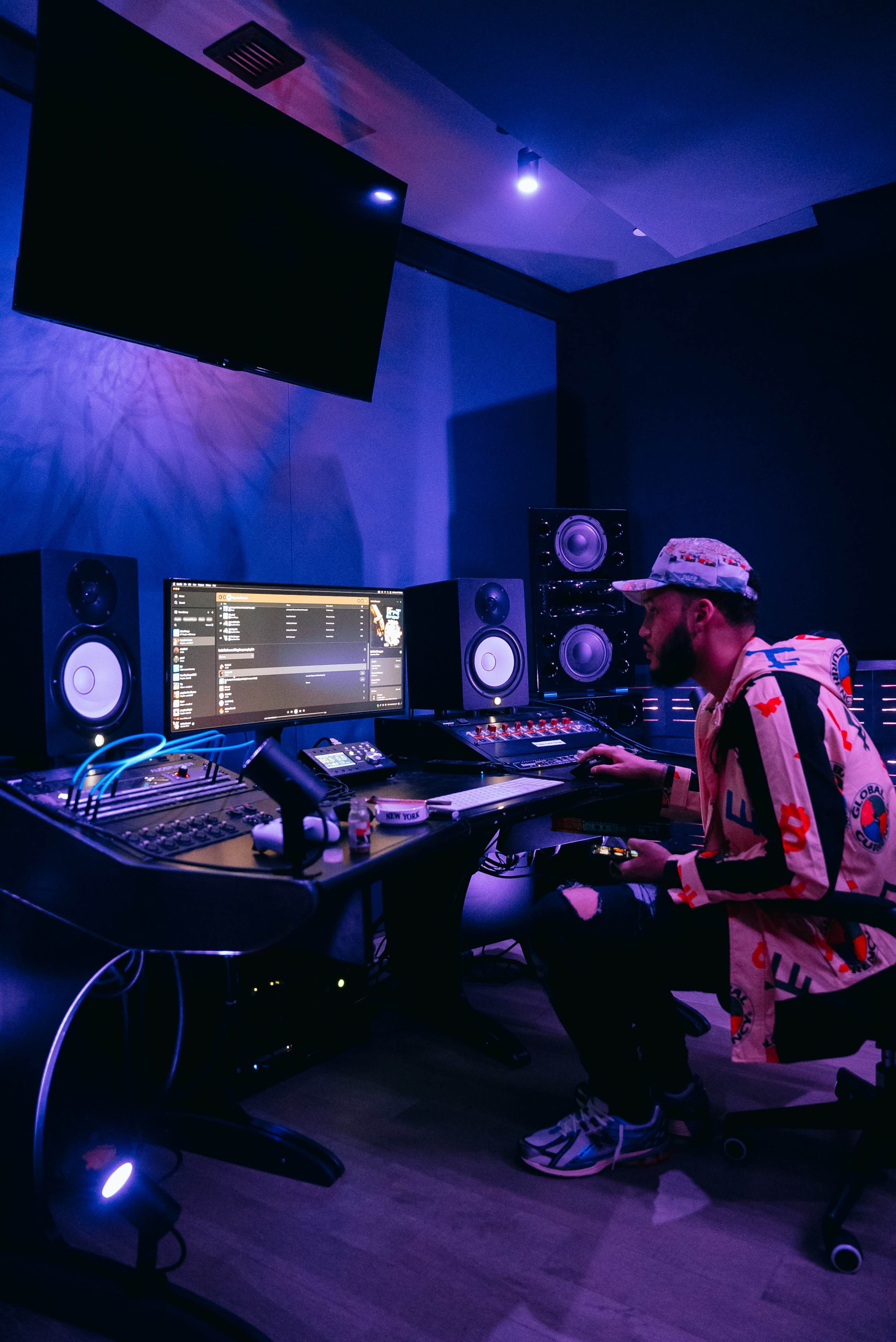 A musician sits at a mixing desk in a dimly lit recording studio, surrounded by speakers and a computer displaying audio tracks.