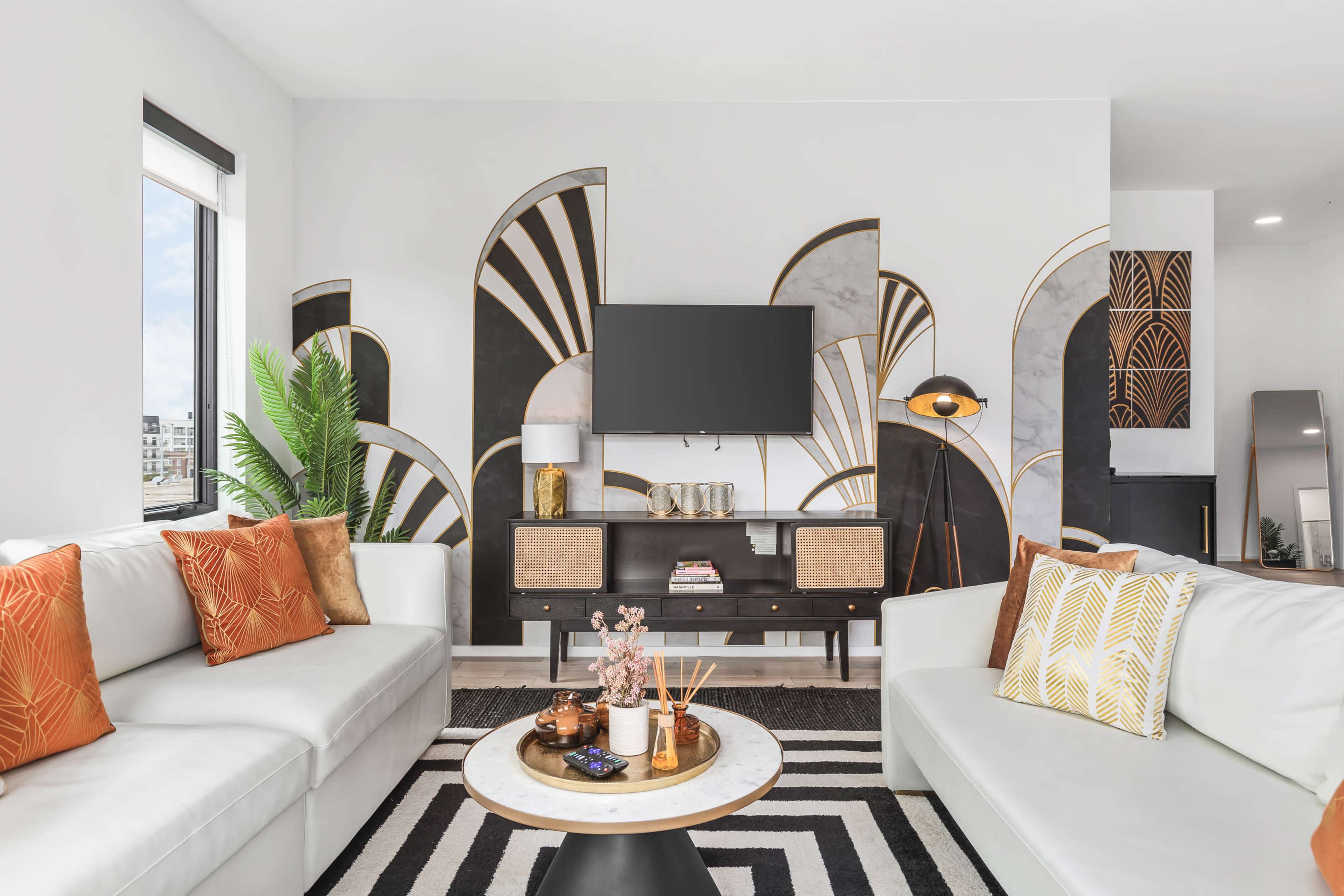 A modern living room features a black and white mural behind a stylish TV unit, with a round coffee table at the center and plush sofas adorned with orange and gold cushions.