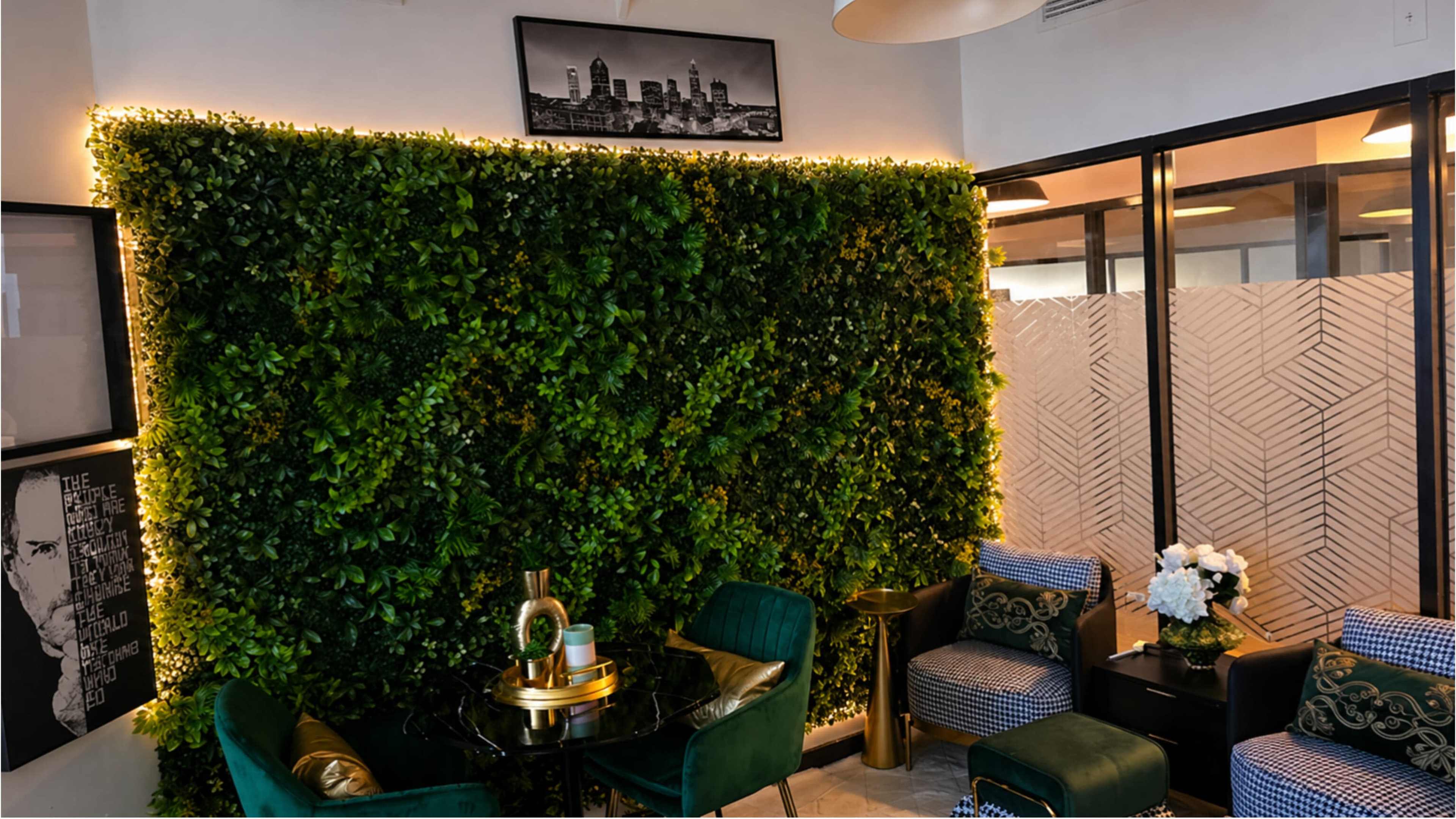 The image shows an indoor space featuring a green wall with artificial foliage, illuminated by soft lighting, alongside stylish furniture and decorative elements.