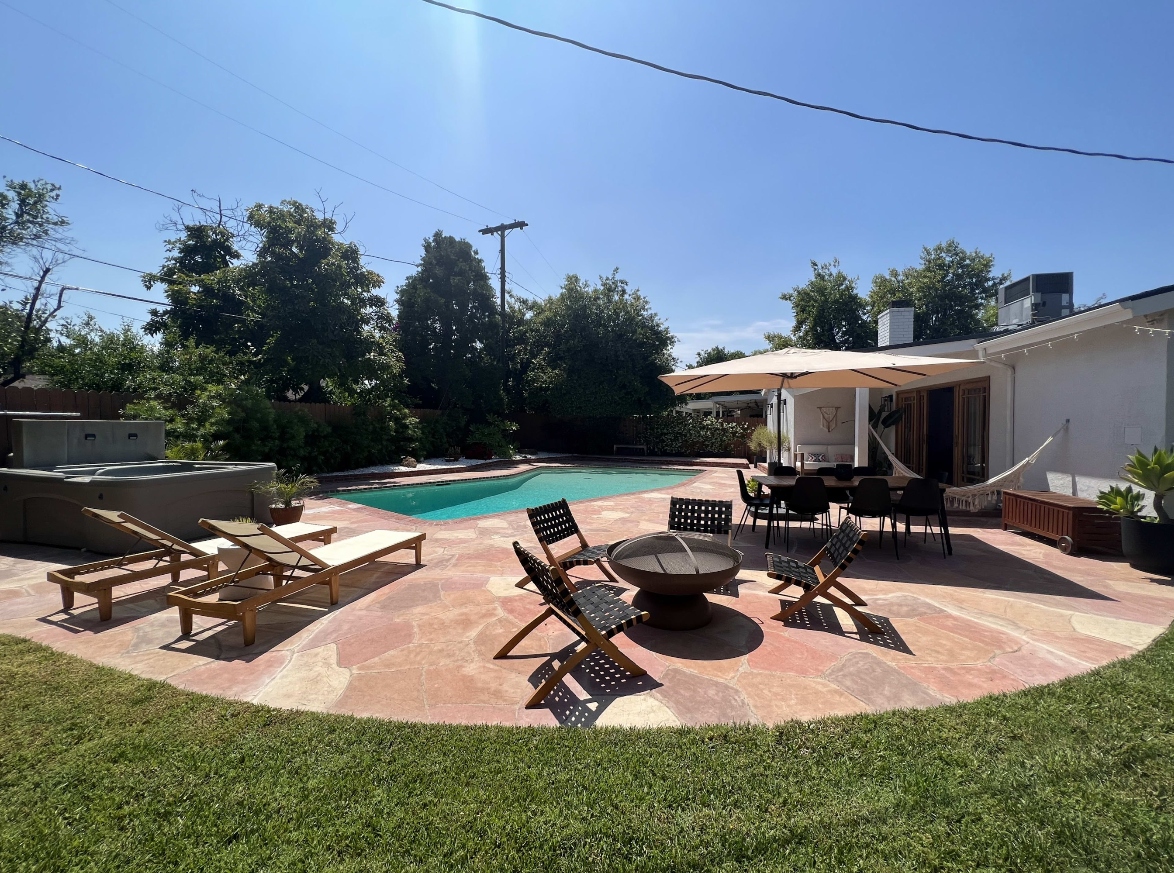 The image shows a backyard with a swimming pool, wooden lounge chairs, a fire pit, and a shaded dining area.