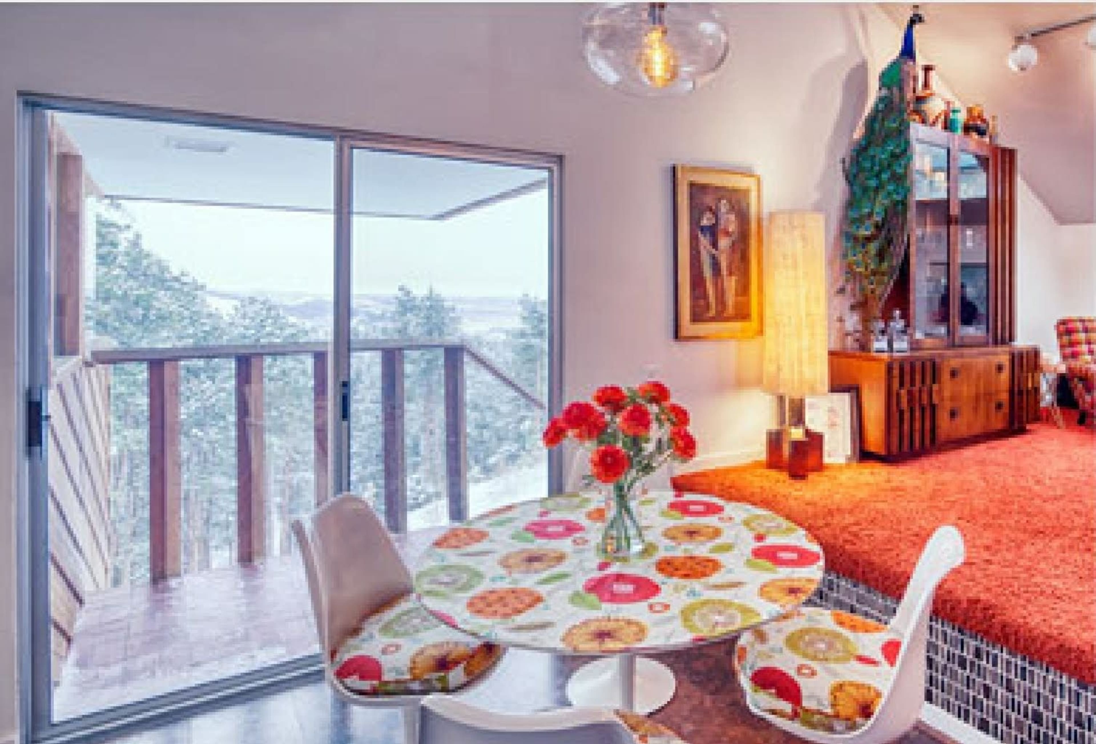 A cozy dining area features a round table with patterned seating, a vase of flowers, and large windows overlooking a scenic outdoor view.