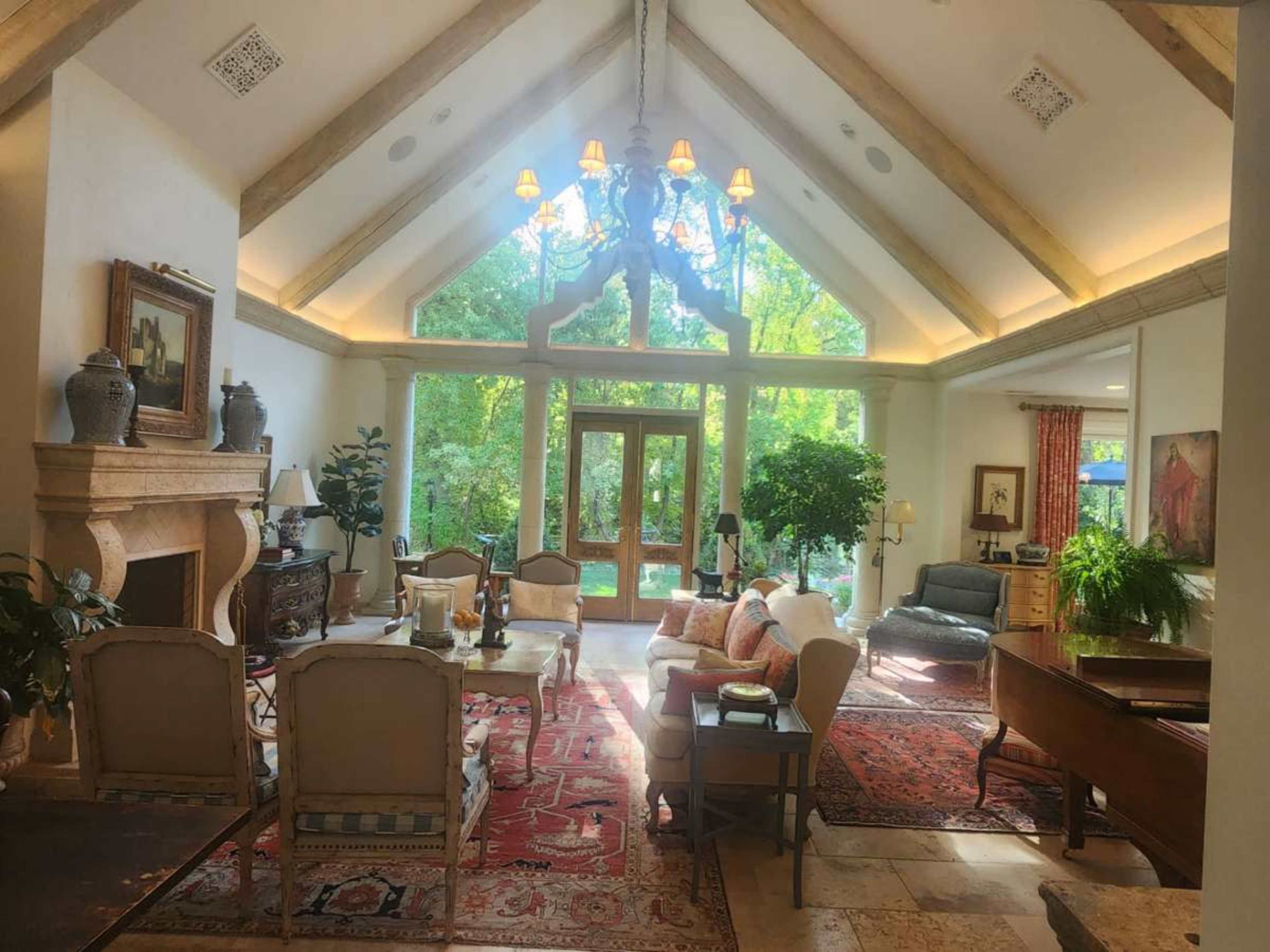 The image shows a spacious living room with tall ceilings, large windows letting in natural light, and a mix of comfortable furniture, including a sofa and chairs, arranged around a patterned area rug.
