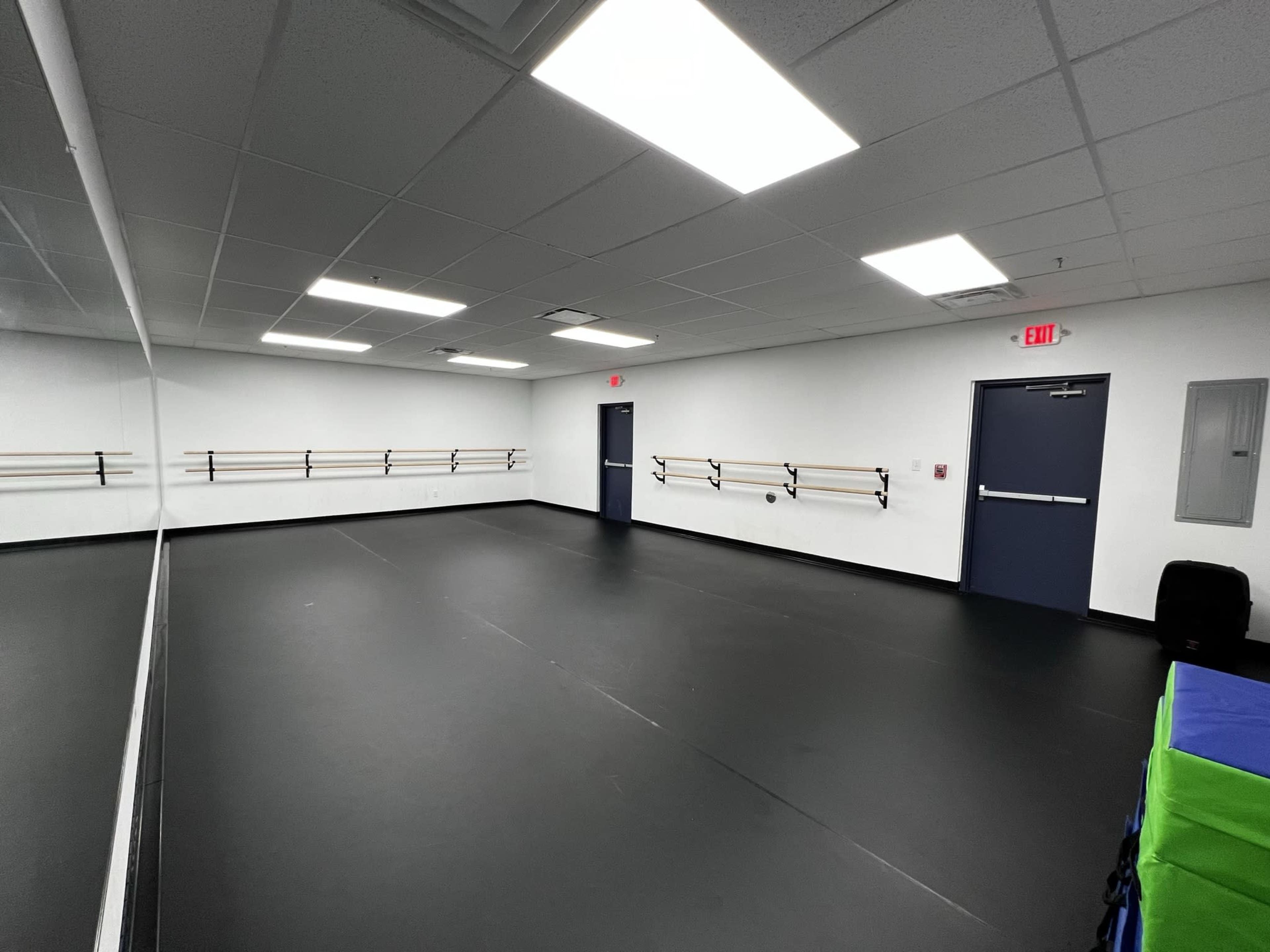 A spacious dance studio with black flooring, mirror walls, and wooden ballet bars mounted on the walls.