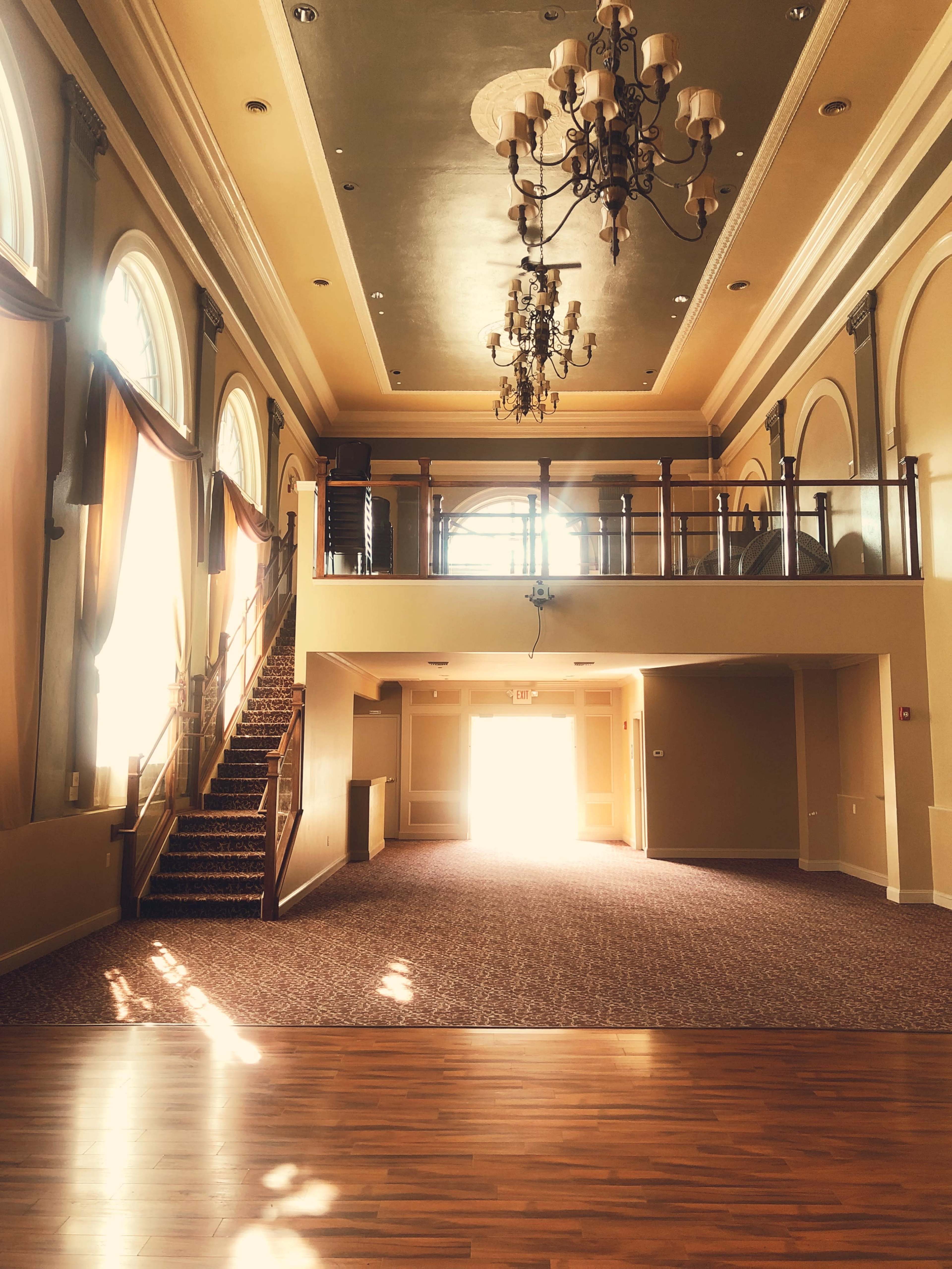 The spacious interior features large arched windows, a staircase leading to a balcony, and chandeliers hanging from a high ceiling.