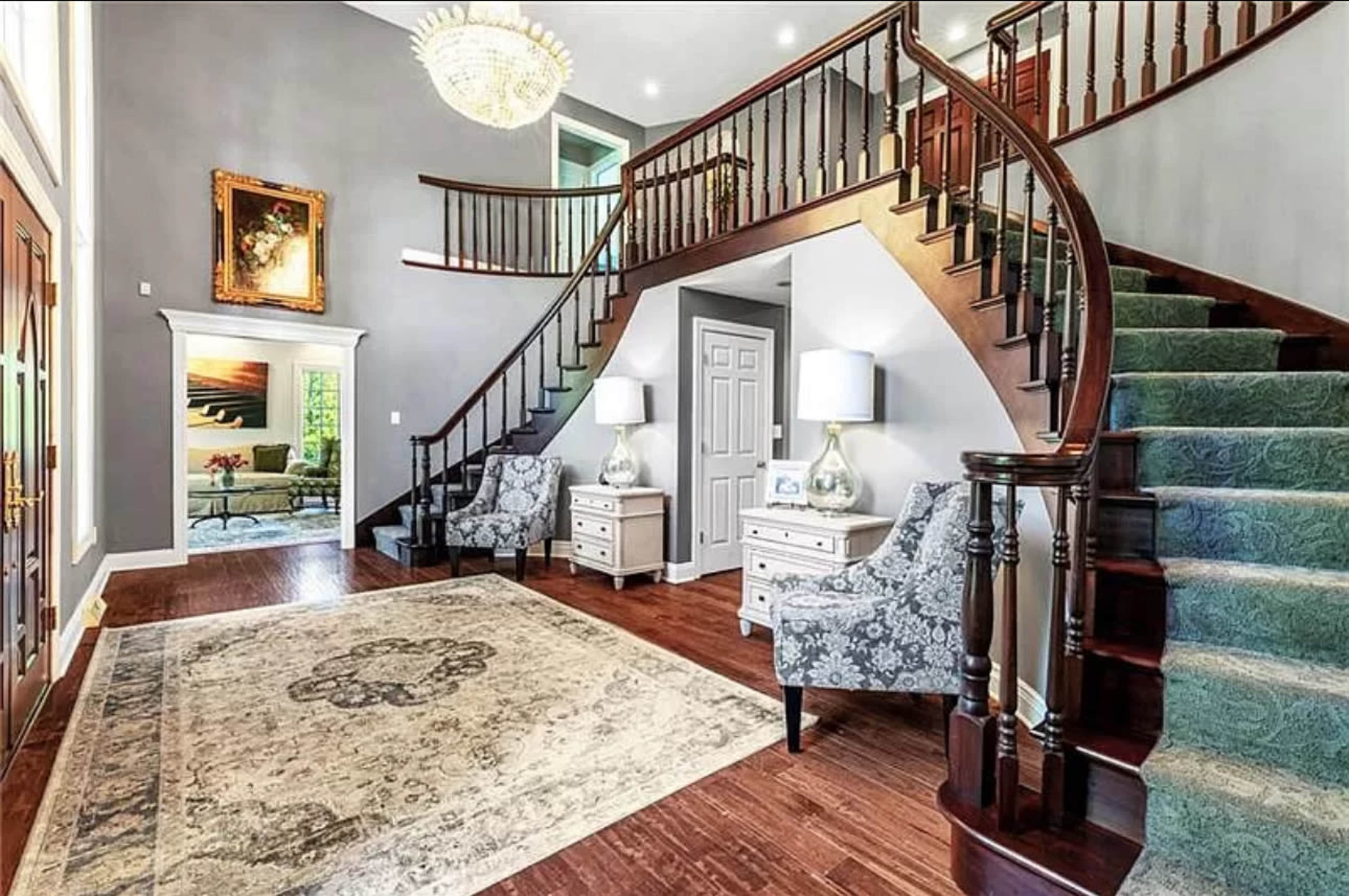 A grand foyer features a curved staircase, two upholstered chairs, a large chandelier, and a decorative area rug on hardwood flooring.
