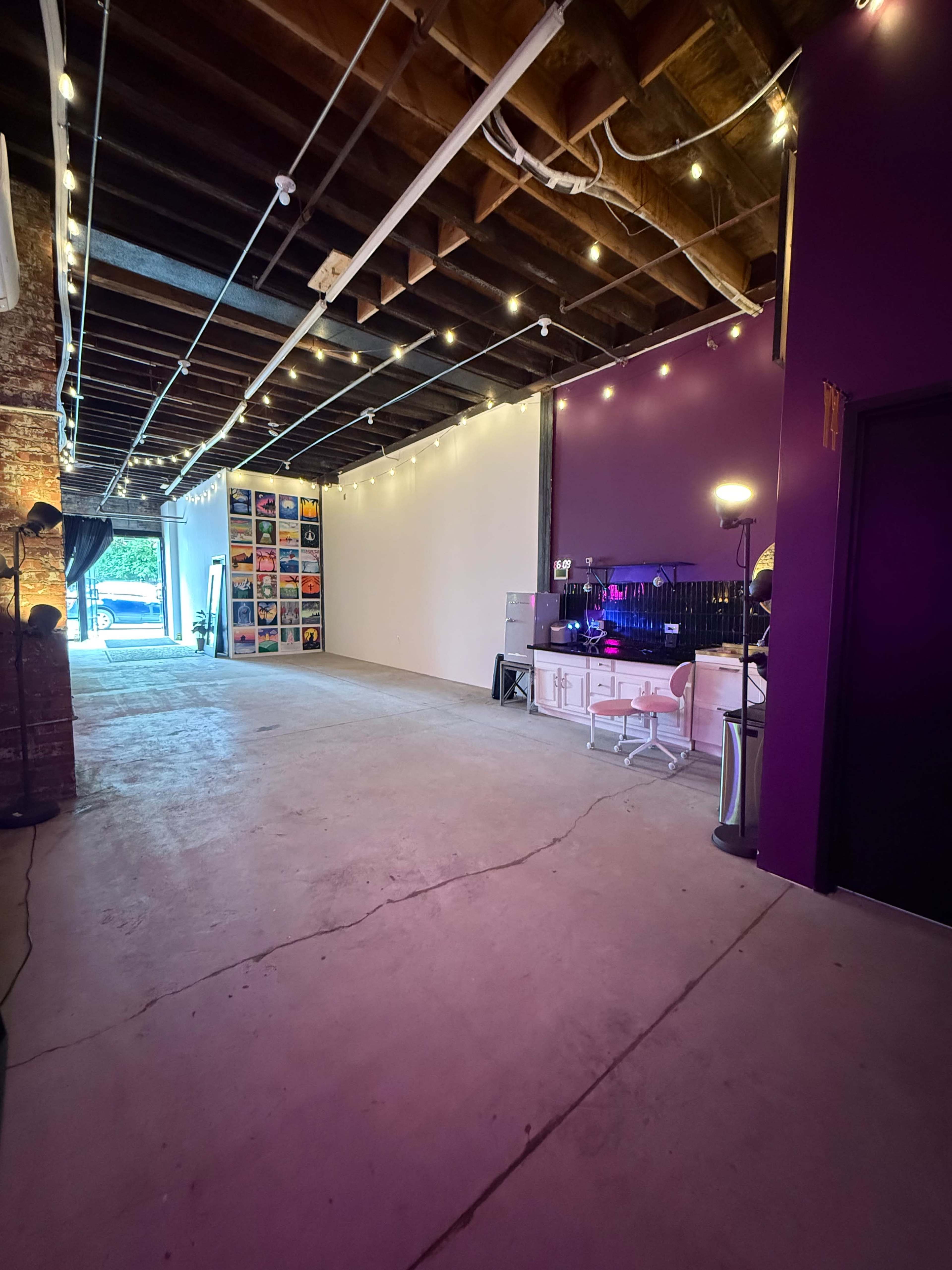 The image shows a spacious, well-lit interior with exposed beams and string lights, featuring a purple wall, art pieces, and an entrance leading outside.
