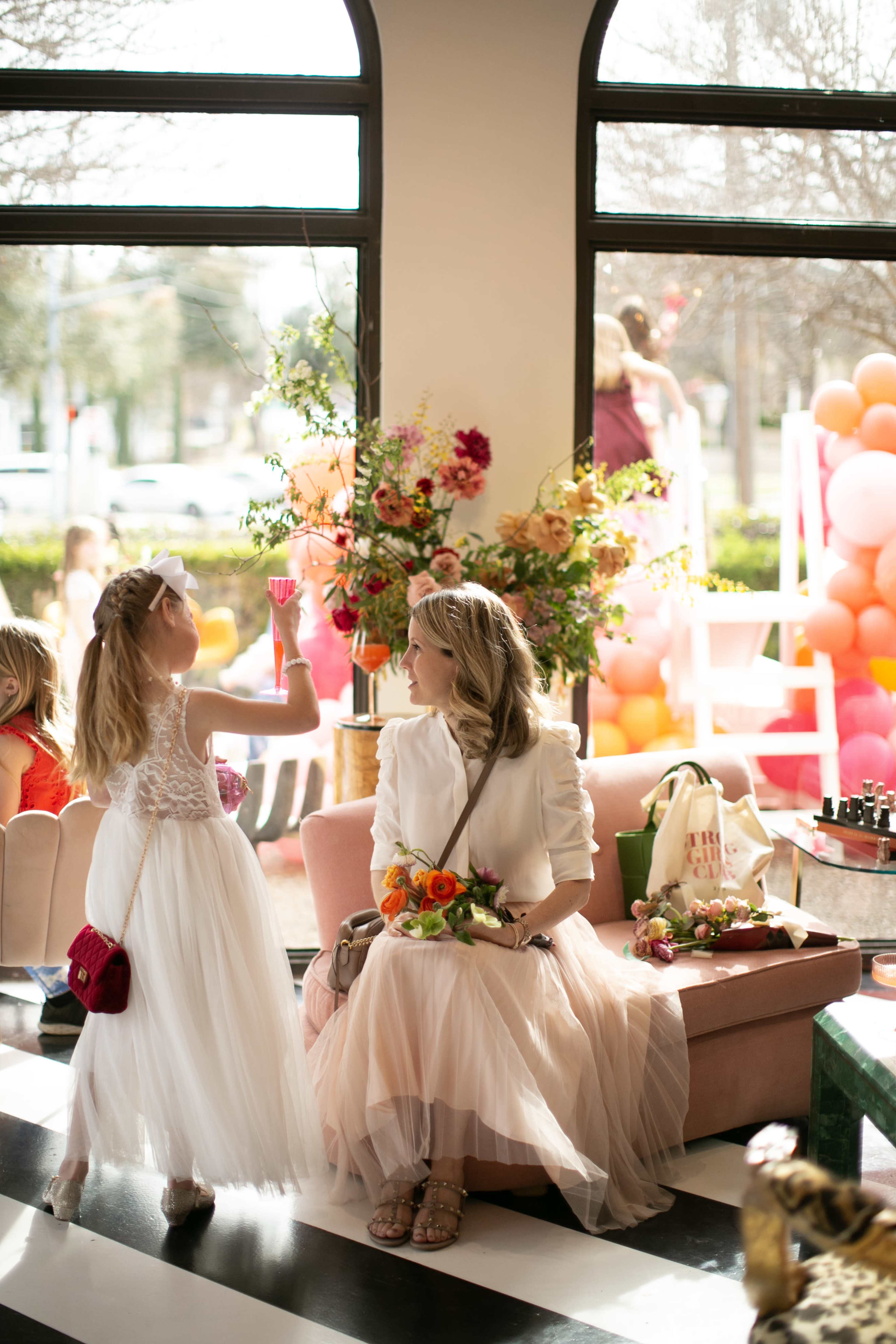 A woman sits on a pink sofa in a brightly lit space adorned with floral arrangements, while a young girl in a white dress holds a small glass near her.