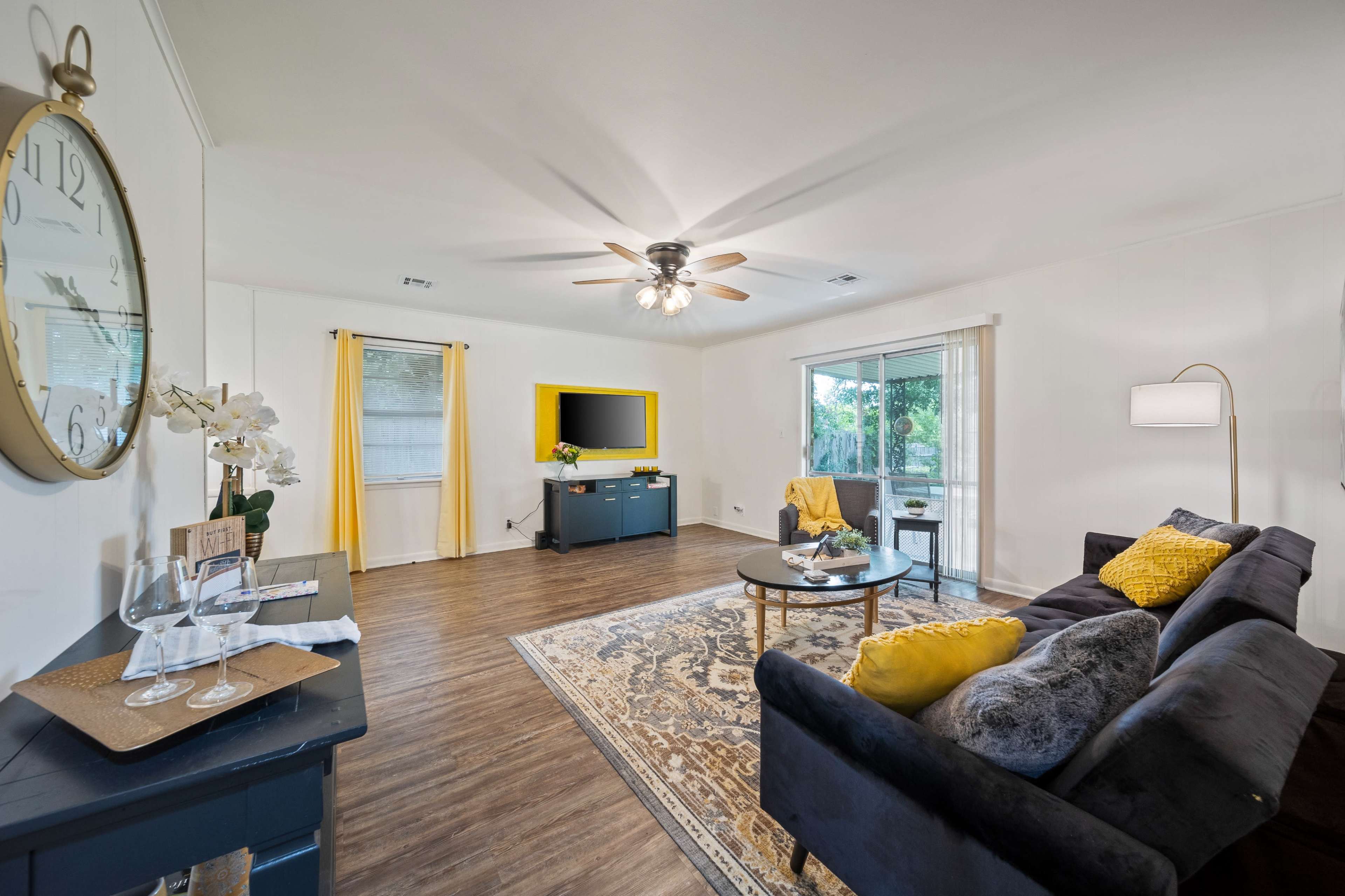 A modern living room features a dark sofa, a round coffee table, and a television mounted on the wall, with yellow accents and large windows revealing an outdoor view.