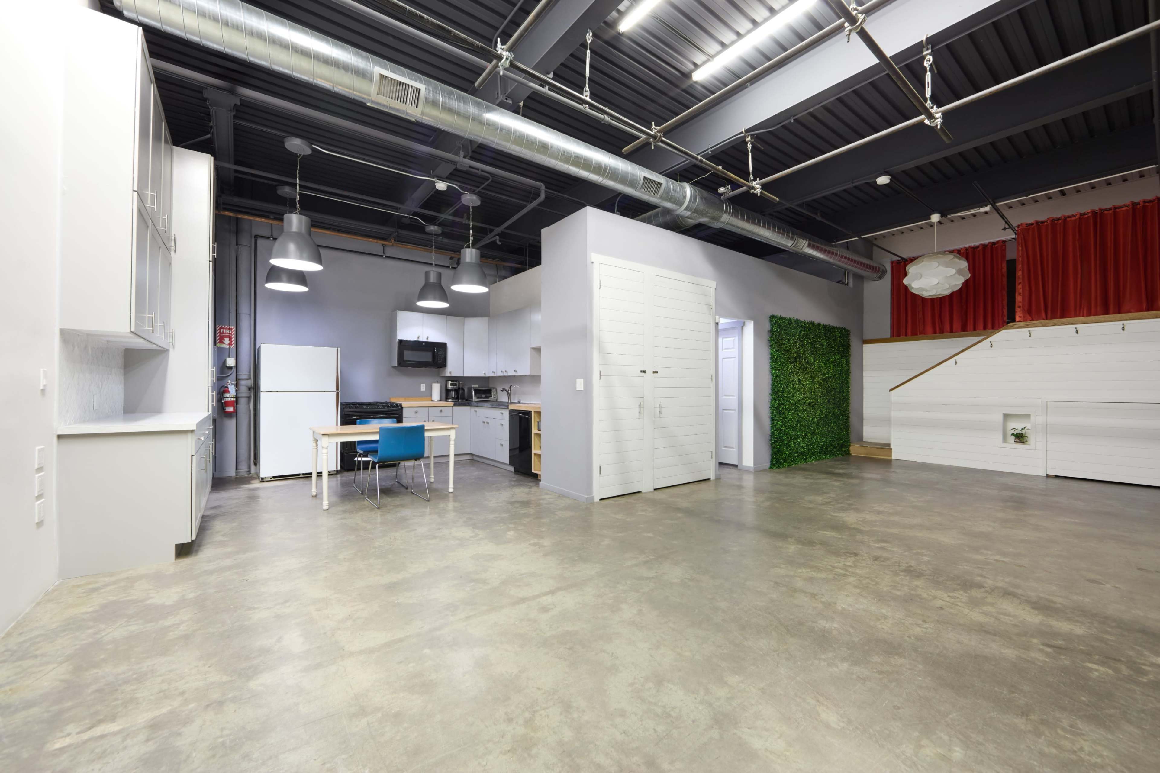 The image shows a spacious, modern interior with a kitchen area, dining table, and lounge space, featuring concrete floors and industrial-style lighting.
