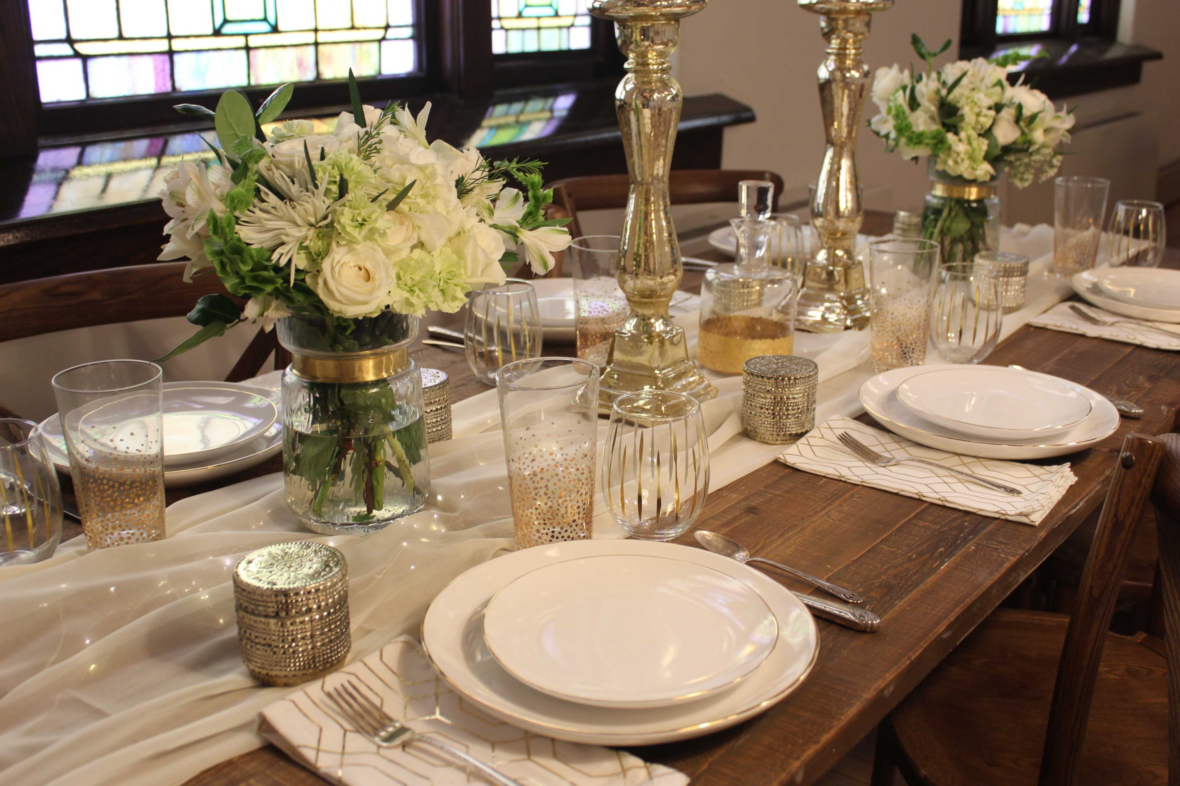 A dining table is set with white plates, floral centerpieces, glassware, and lit candles, all arranged on a white table runner.