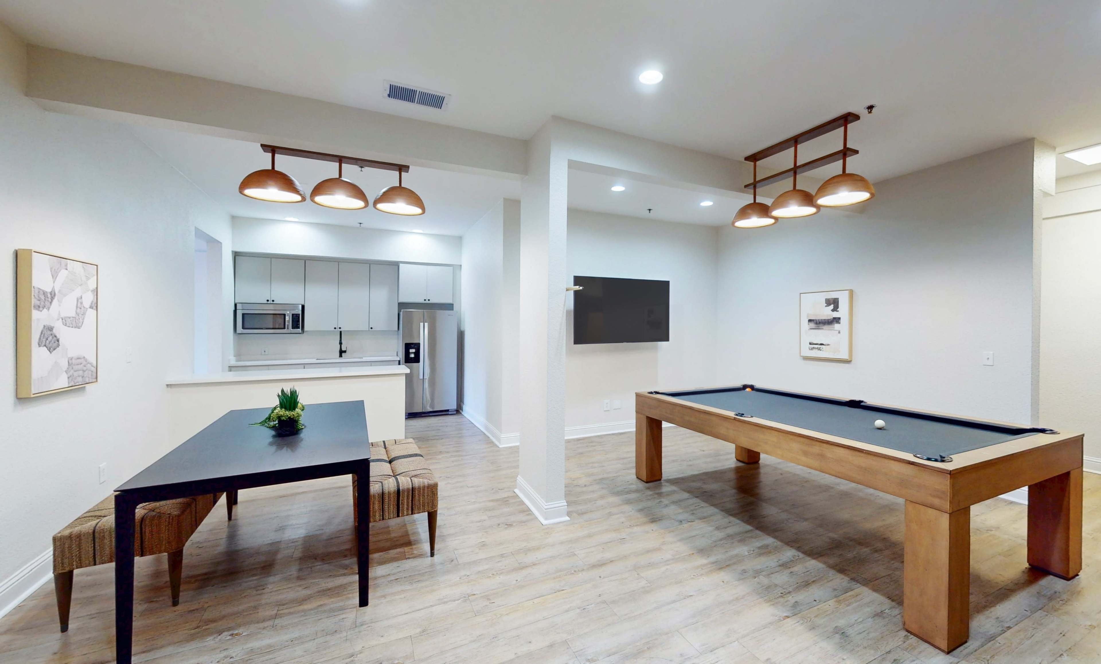 The image shows a spacious game room featuring a billiards table, a dining area with a table and benches, and a kitchen area in the background.