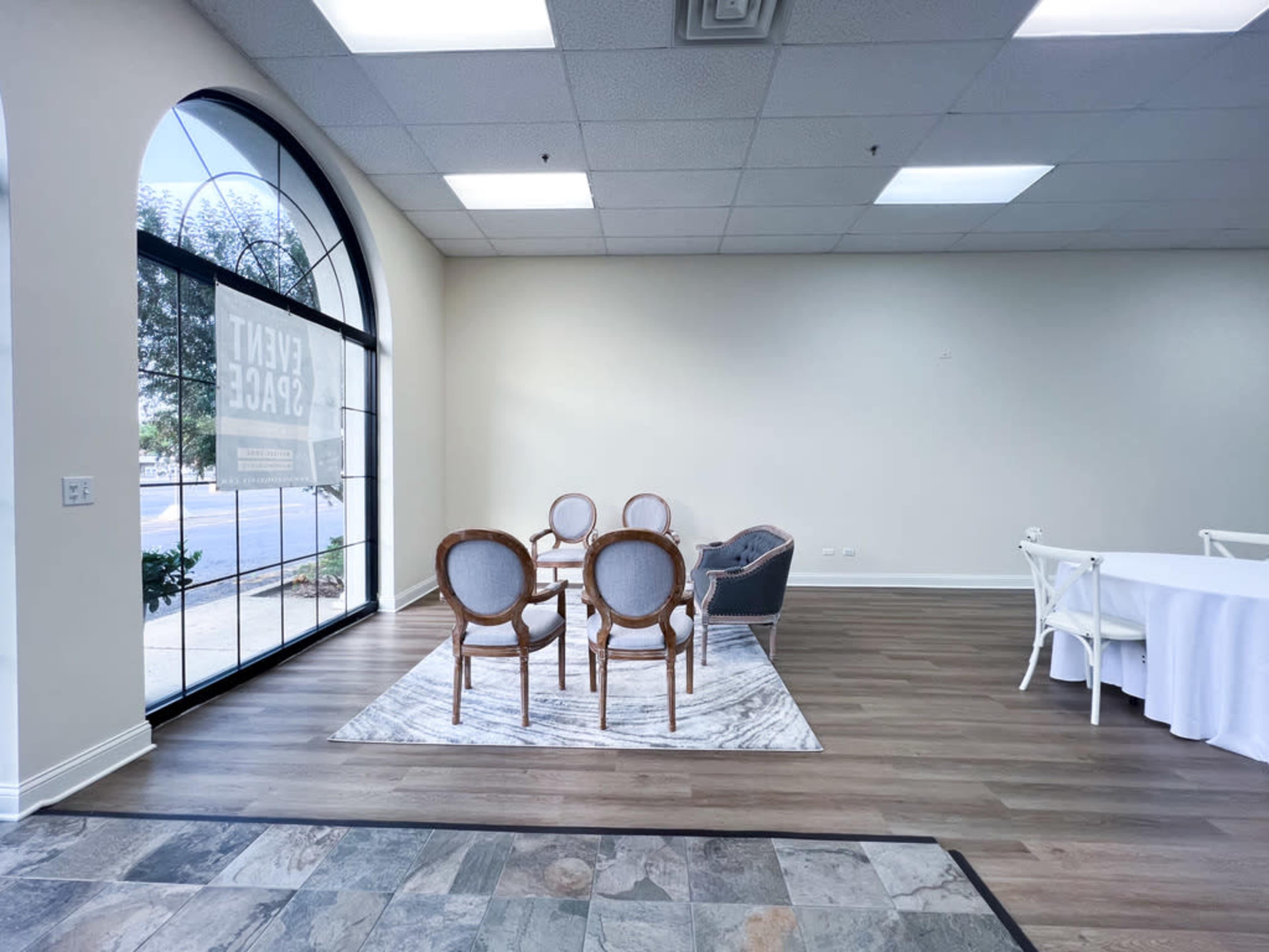 A spacious room with a large window, a seating area featuring a round table and four chairs on a rug, and a partially visible dining setup in the background.