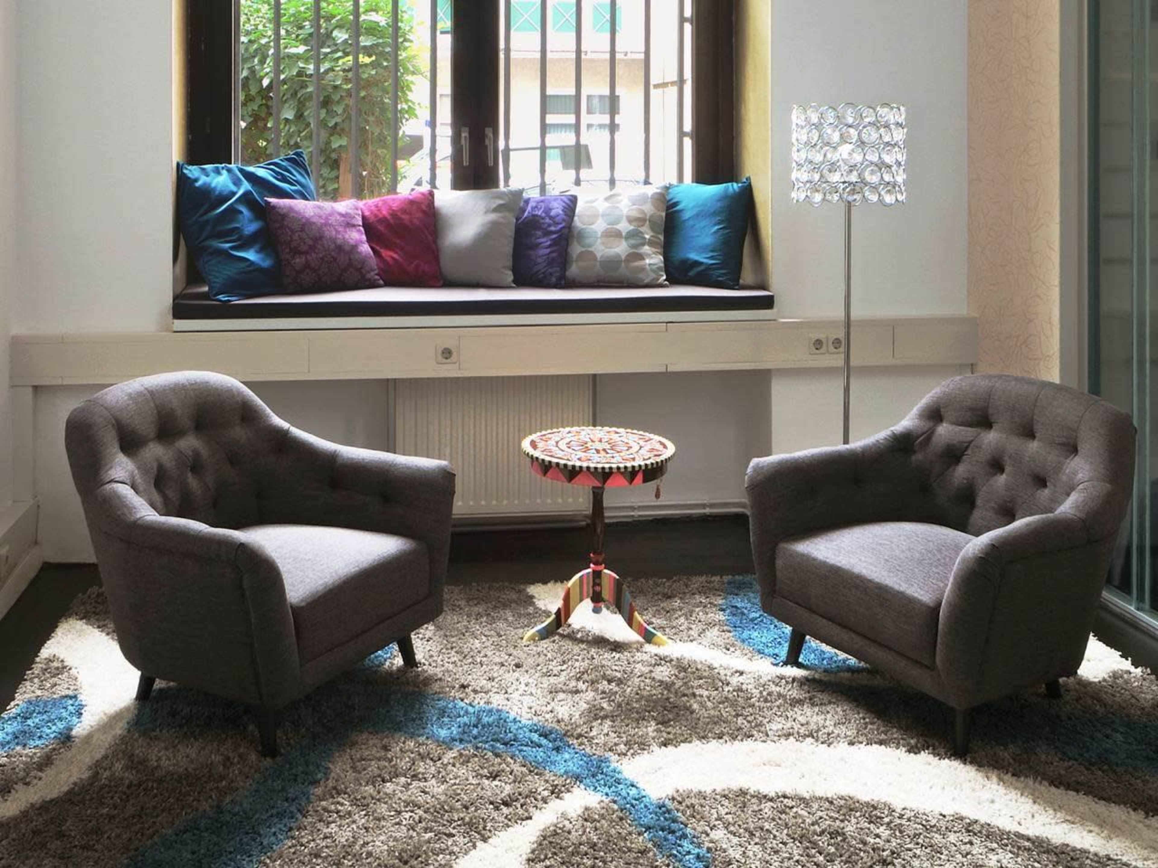 Two gray armchairs face a small, ornate table on a patterned rug, with a cushion-filled window seat and a floor lamp in the background.