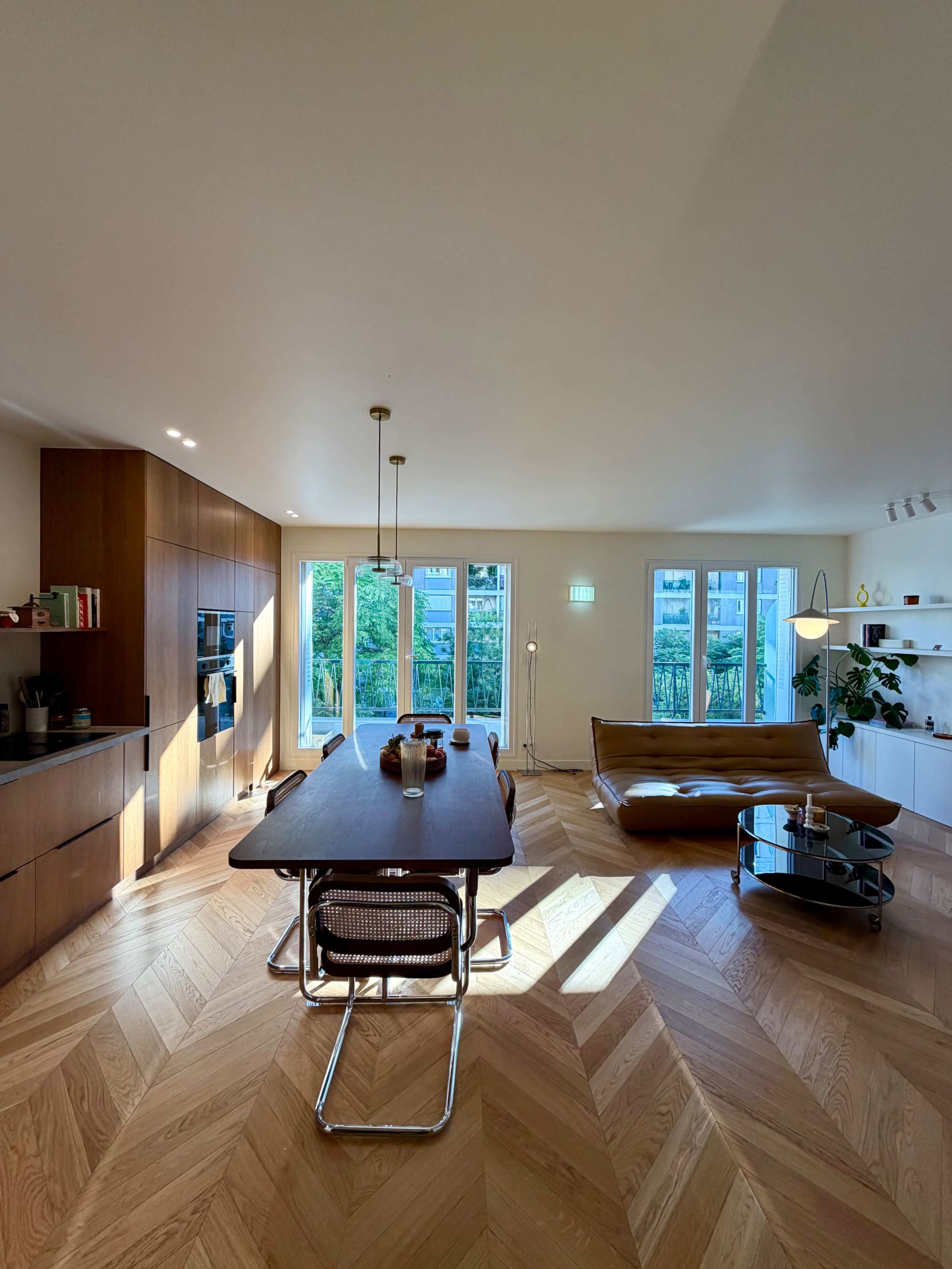 The image shows a modern living space with a dining table, wooden cabinets, large windows letting in natural light, and minimalist decor.