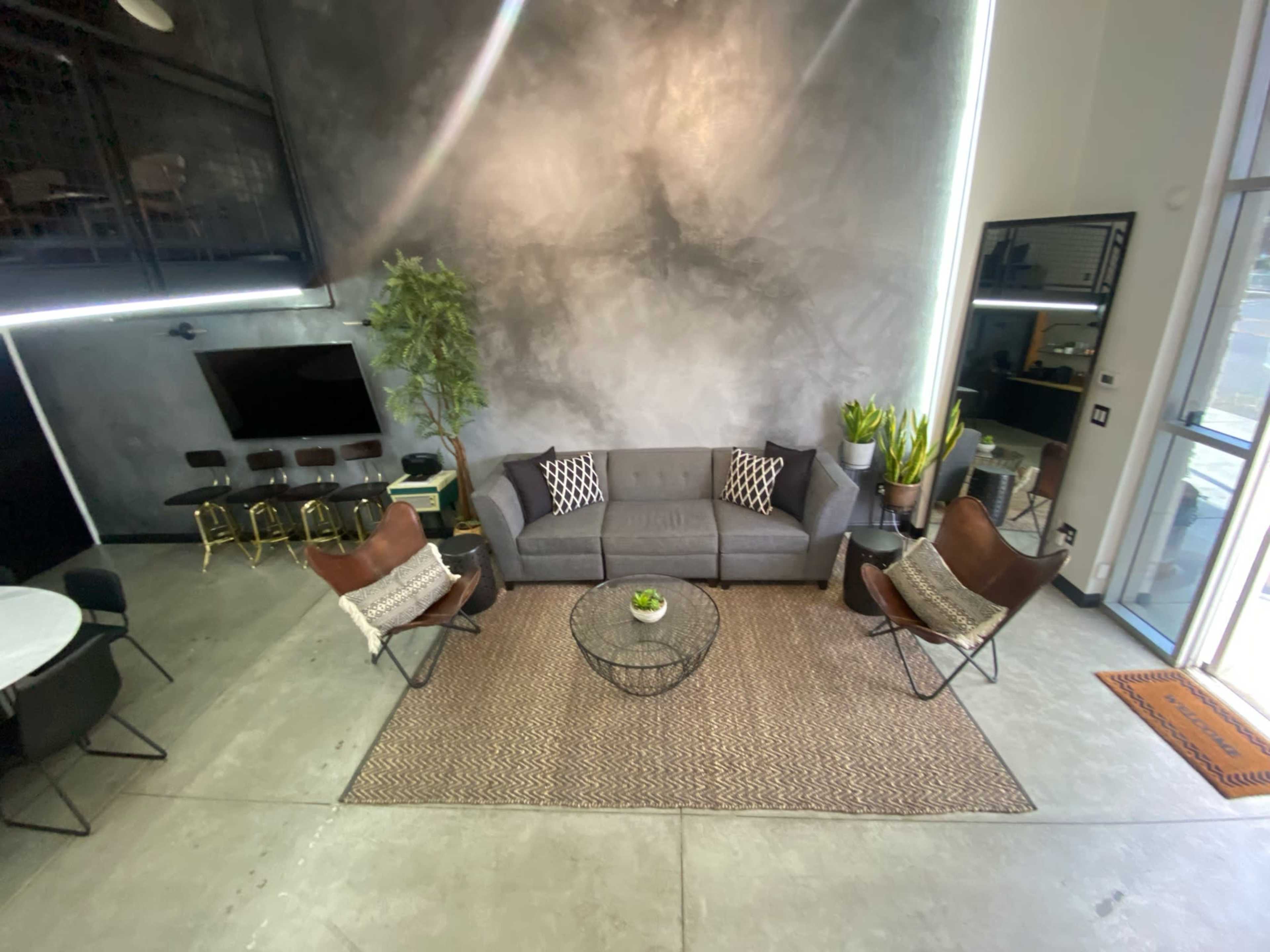 A modern living space with a gray sofa, two brown leather chairs, a round coffee table, and a rug, surrounded by plants and a large mirror.