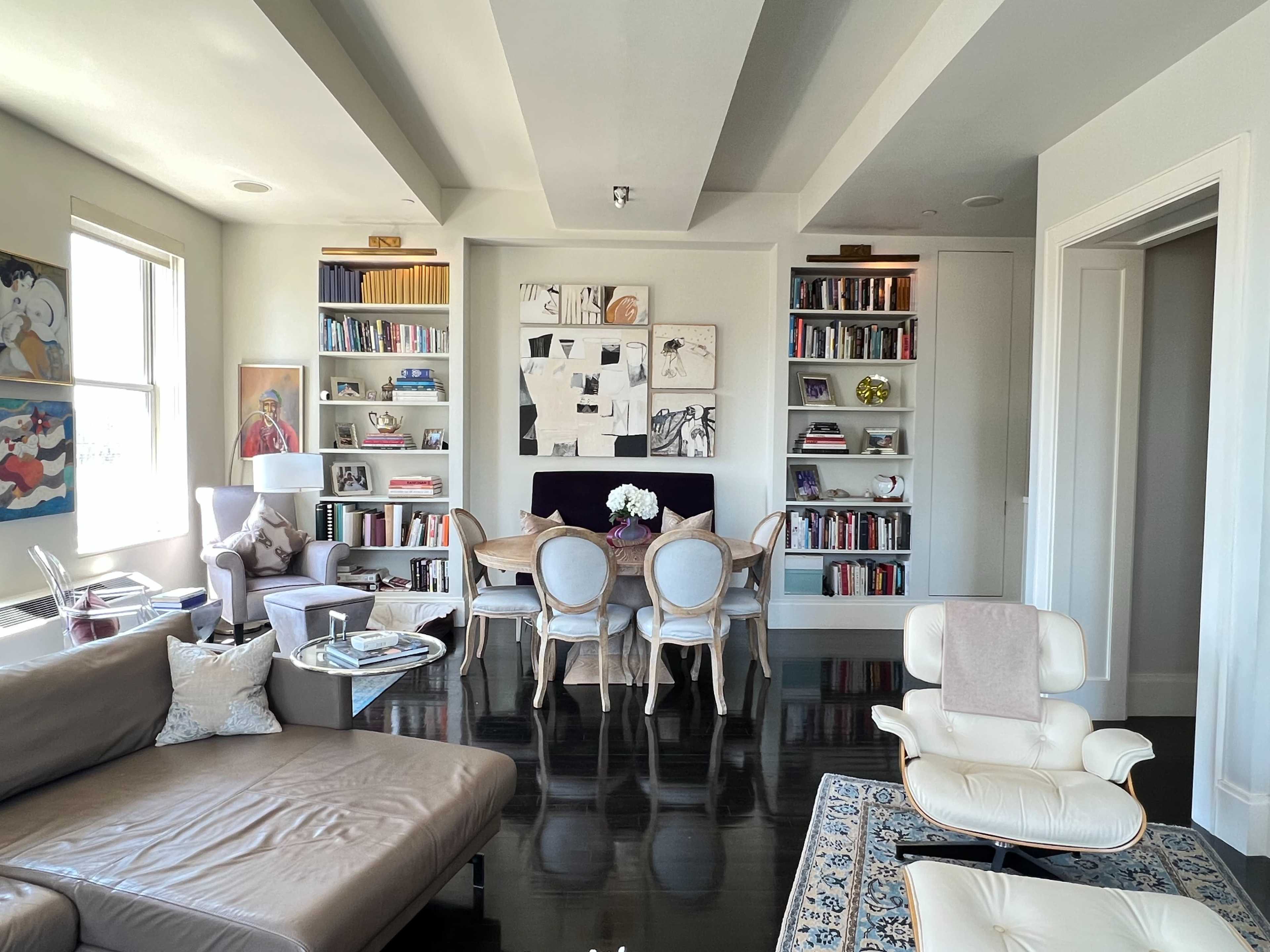 A modern living room features a large sofa, dining area with chairs and a table, bookshelves filled with books, and artwork on the walls.