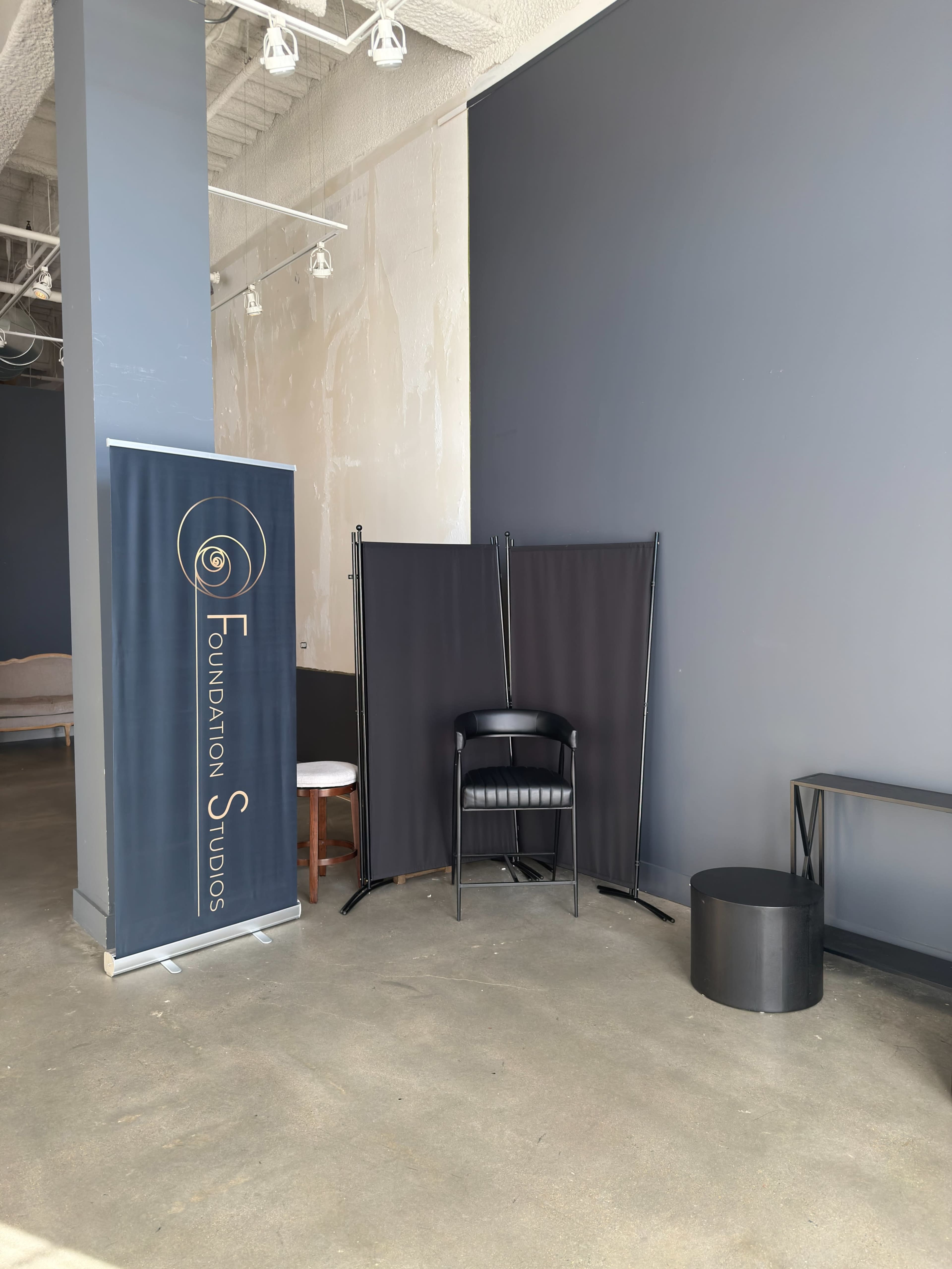 The image shows a corner of a studio space featuring a banner for "Foundation Studios," a folding screen, a black chair, and a small round table.
