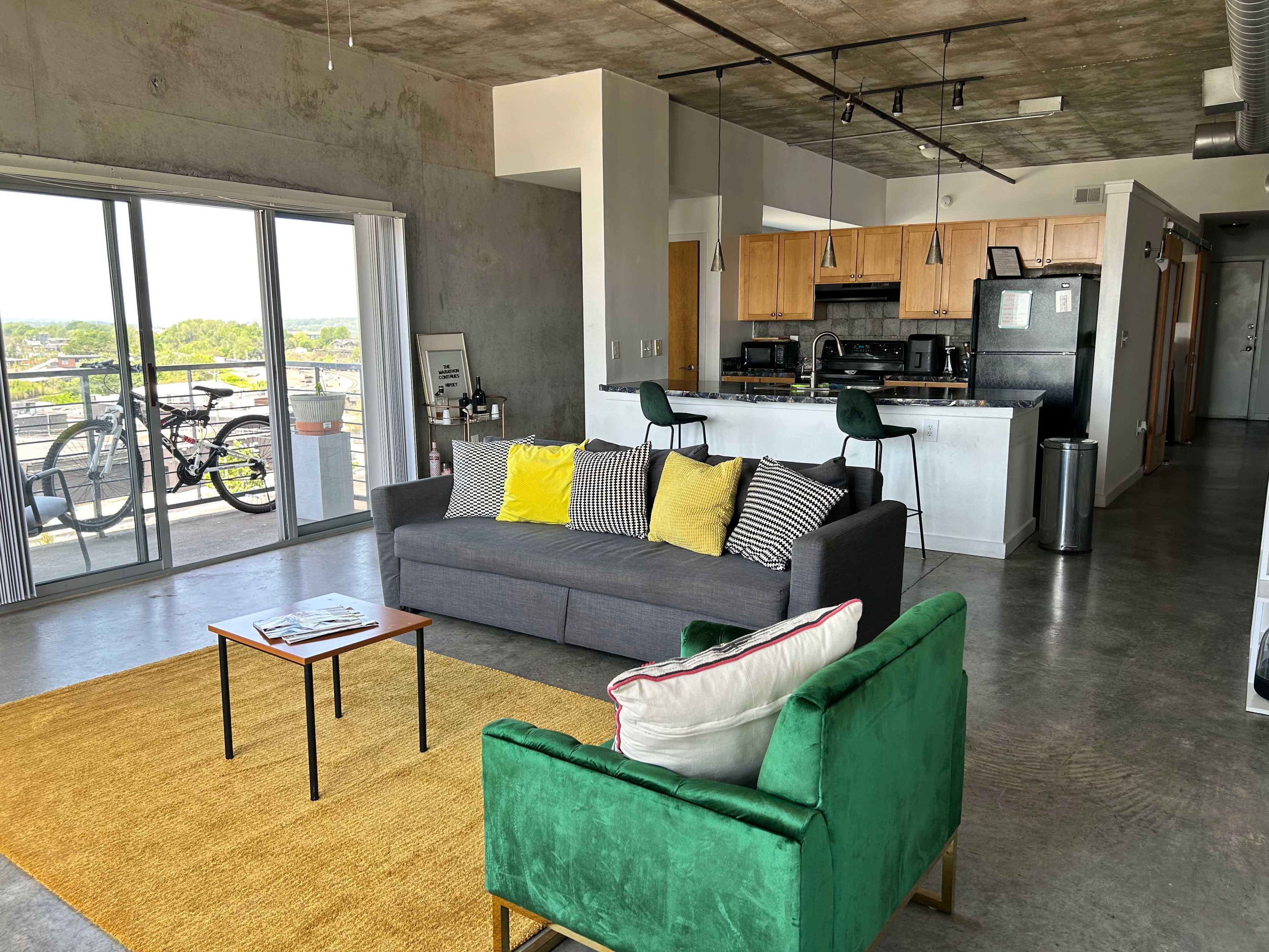 The image shows a modern living space with a gray sofa, a green chair, a small coffee table, and a kitchen area, all bathed in natural light from large windows.