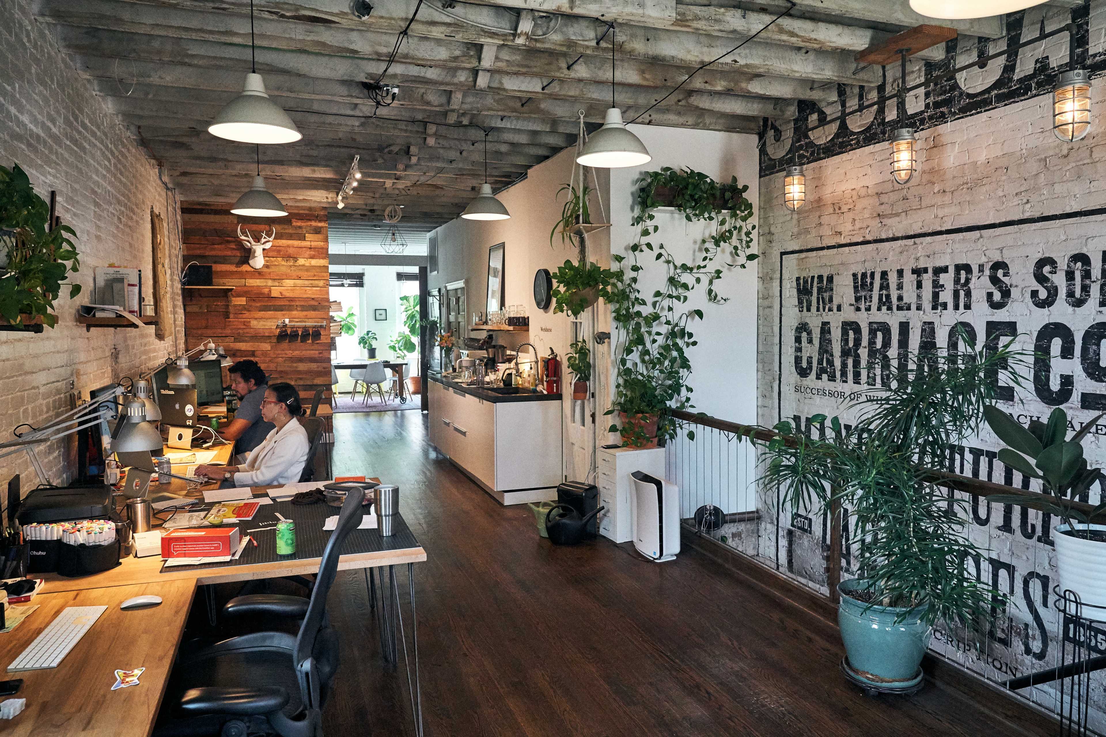 The image shows a spacious office interior with wooden beams, exposed brick walls, and plants, featuring workstations with two individuals seated at desks and a kitchenette area in the background.