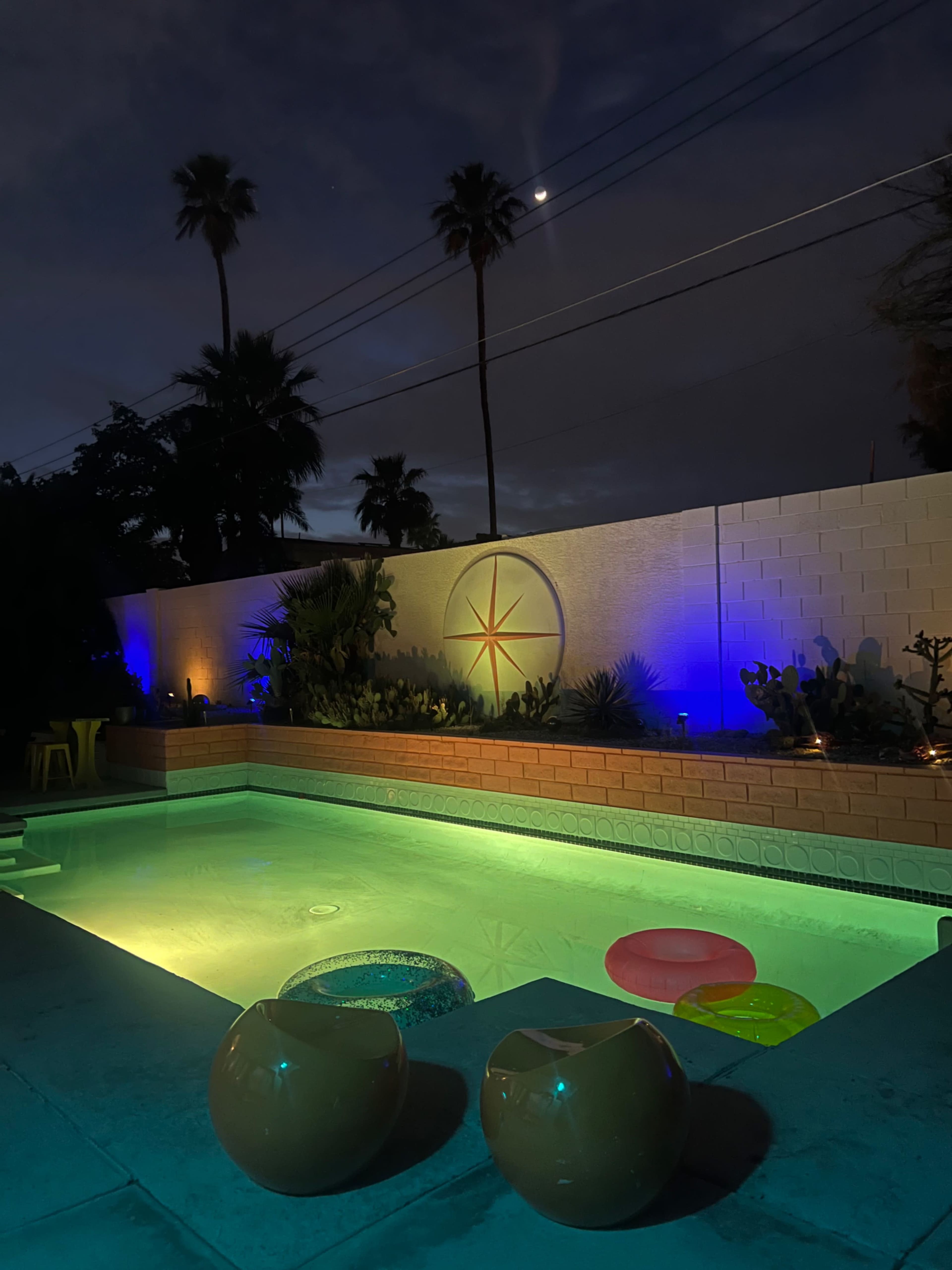 A lit swimming pool with colorful floats and palm trees is set against a night sky illuminated by a crescent moon.