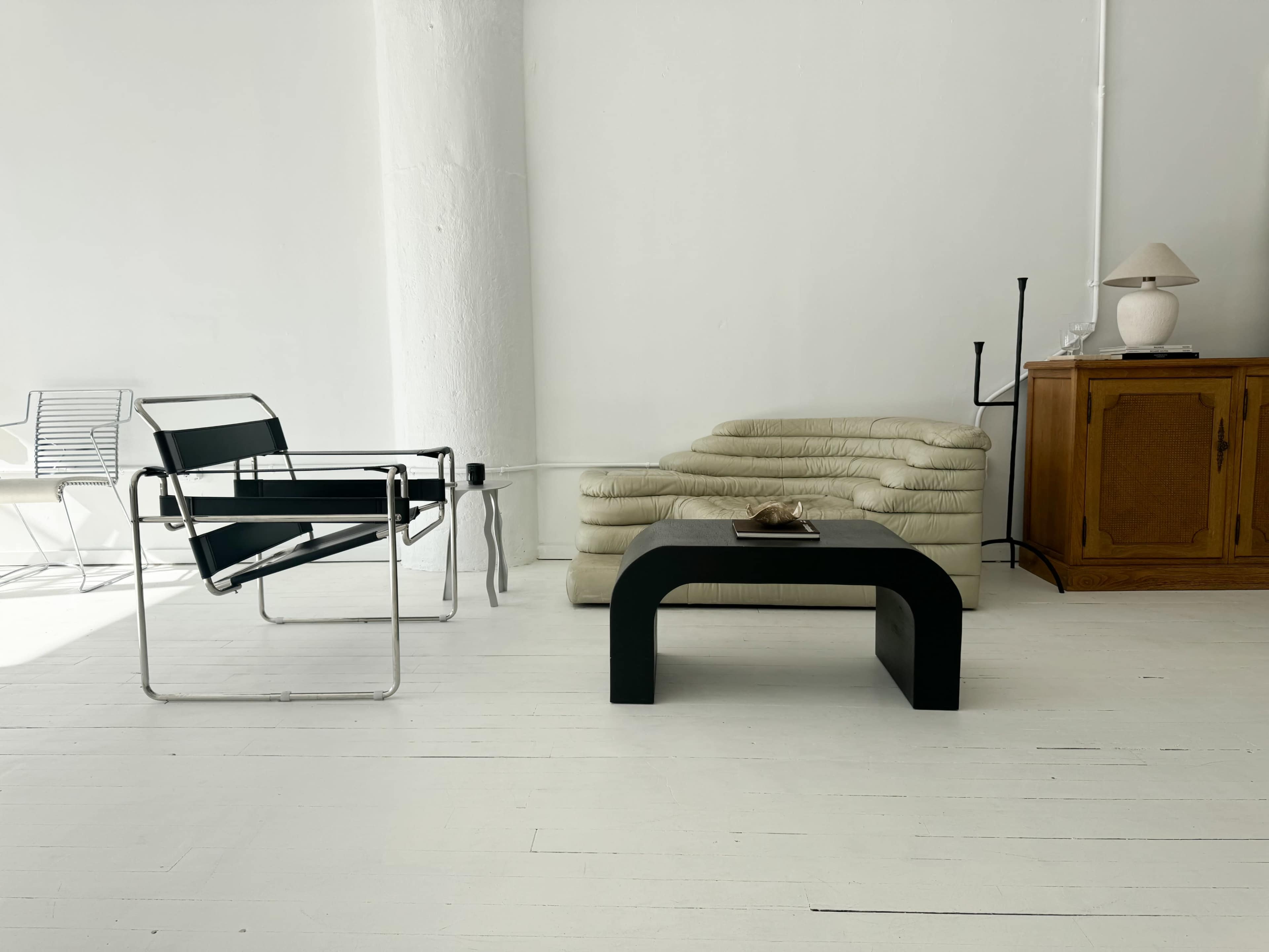 The image shows a minimalistic interior space featuring a black chair, a stack of cream-colored cushions, a black coffee table, and a wooden cabinet with a lamp.