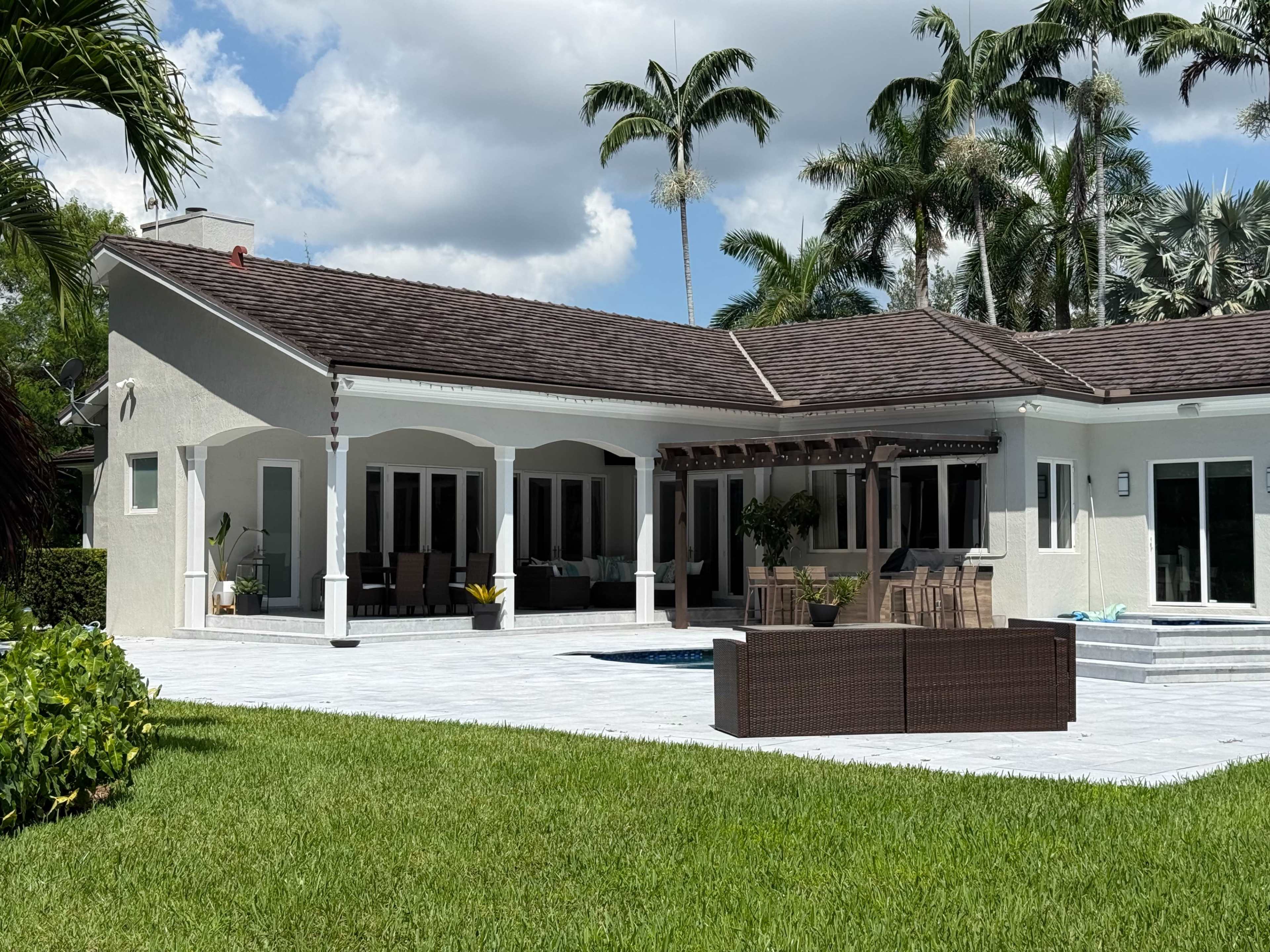 The image shows a modern house with a spacious patio area, palm trees in the background, and a small pool adjacent to the outdoor seating.