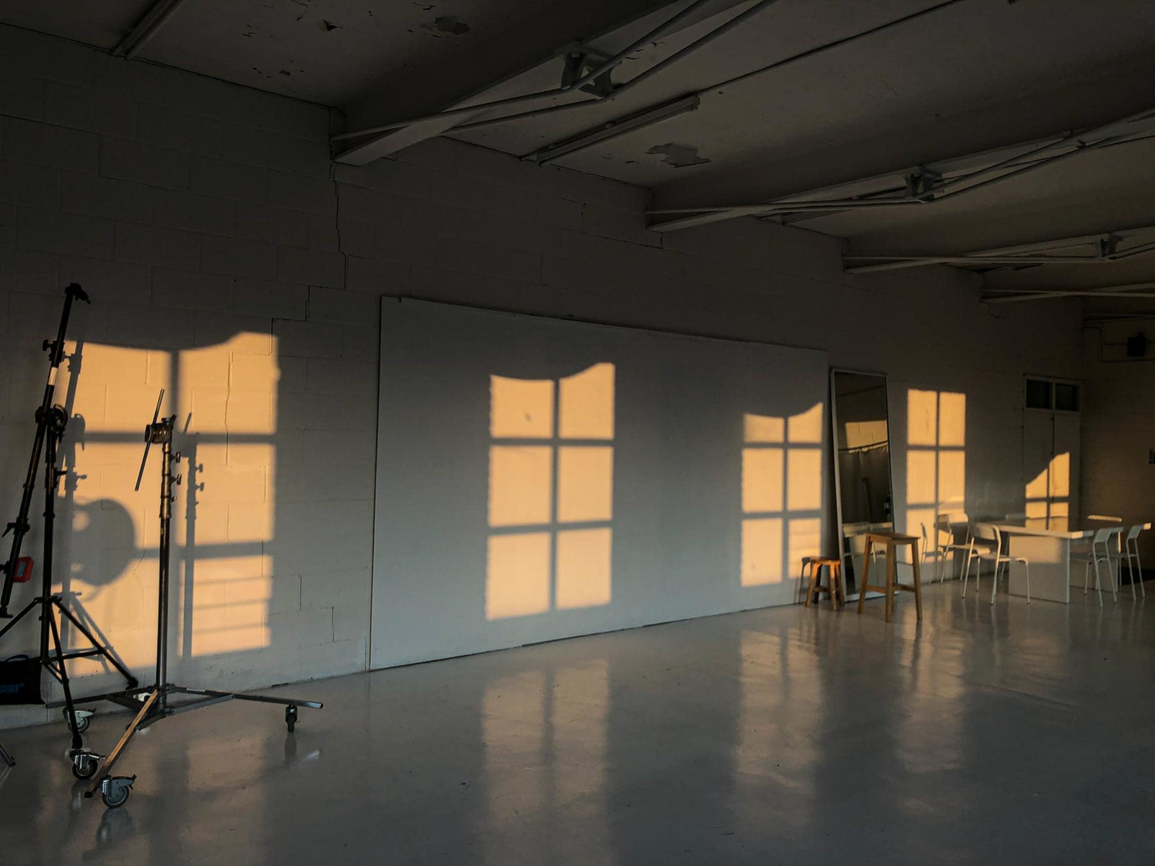 Shadows of a window and furniture are cast on a white wall in a sparsely furnished room with a polished floor.