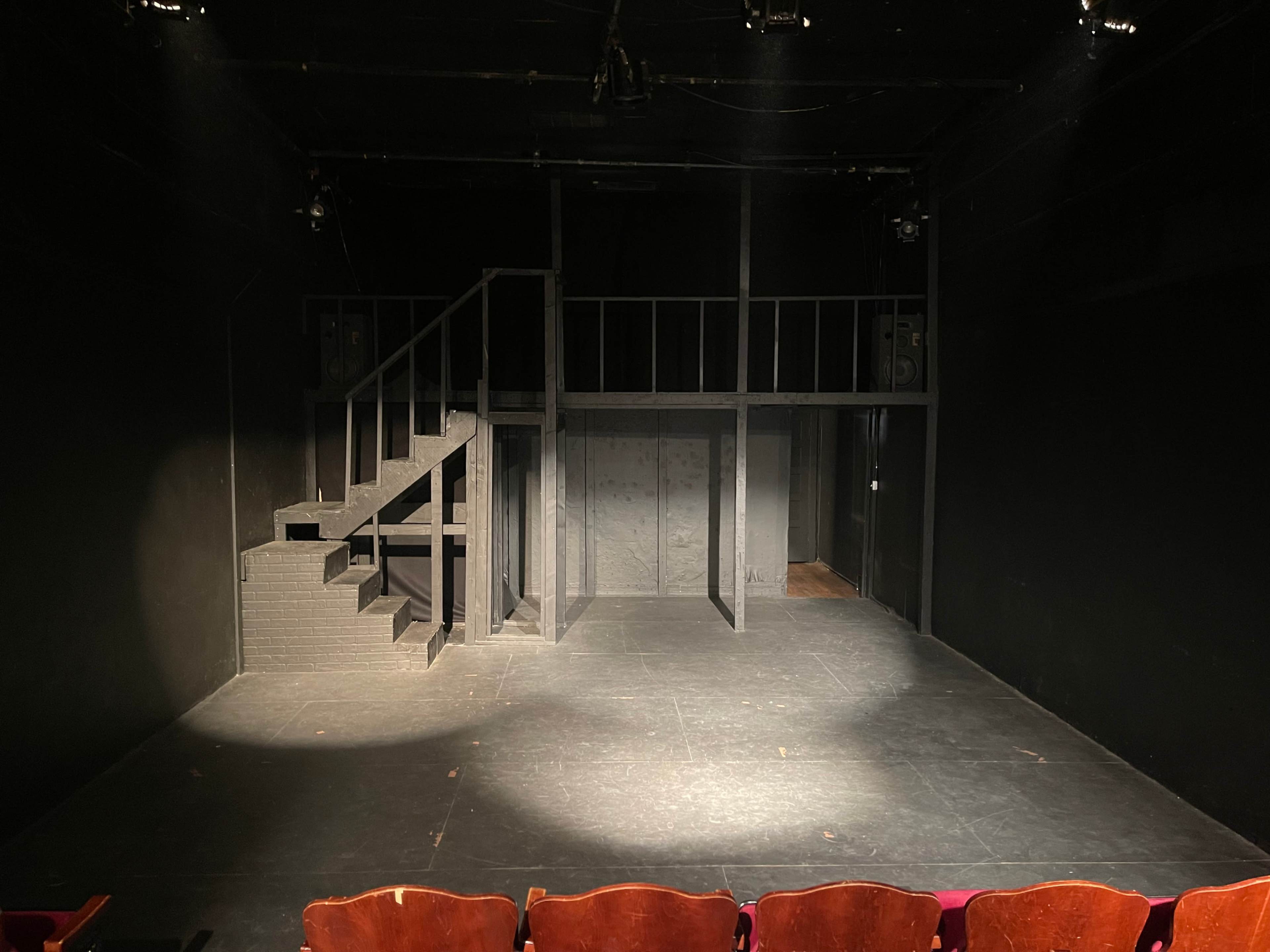 An empty black box theater stage with a staircase on the left and a second-level platform at the back.