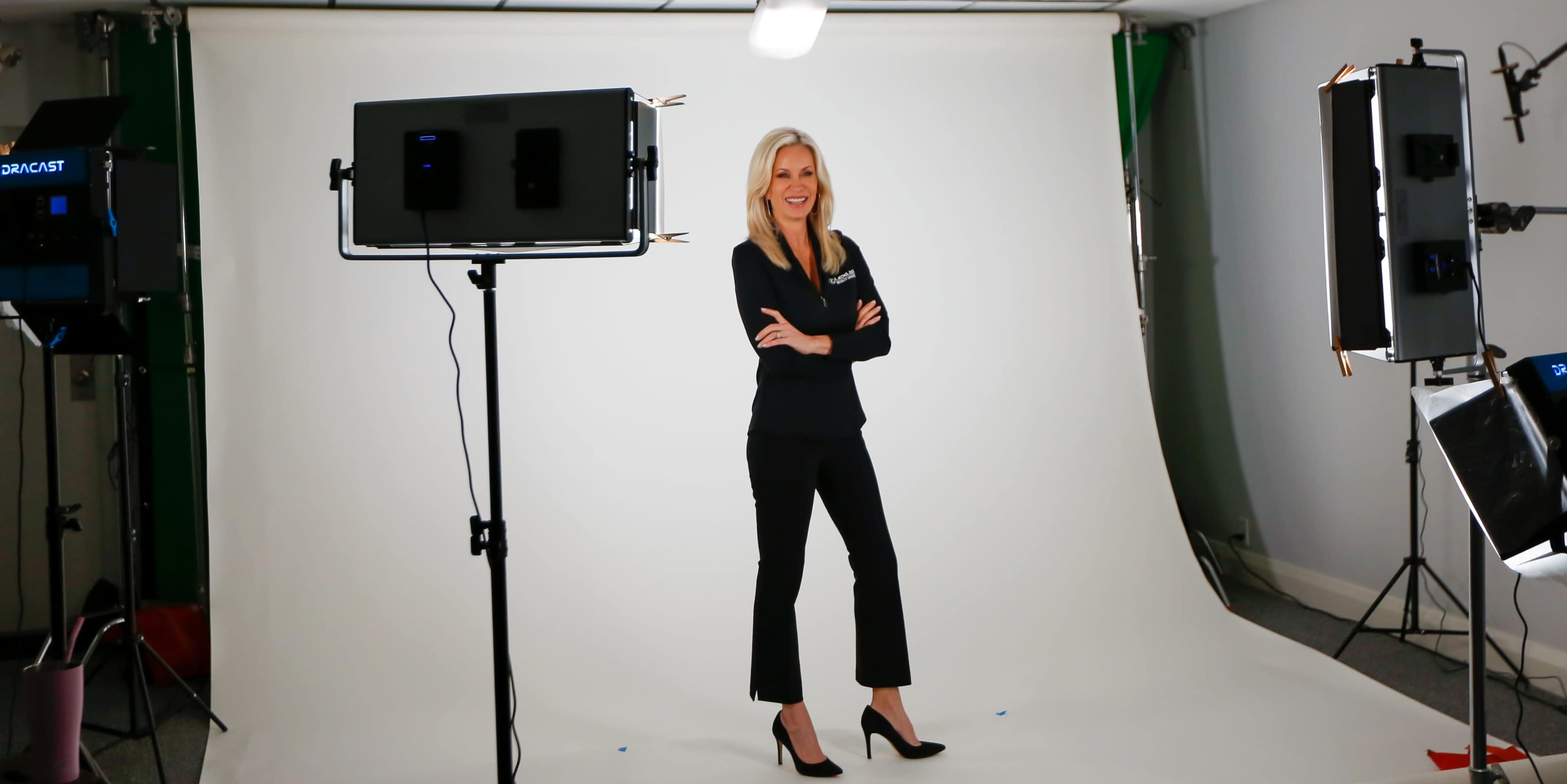 A woman stands with her arms crossed in front of a plain backdrop, surrounded by studio lighting equipment.
