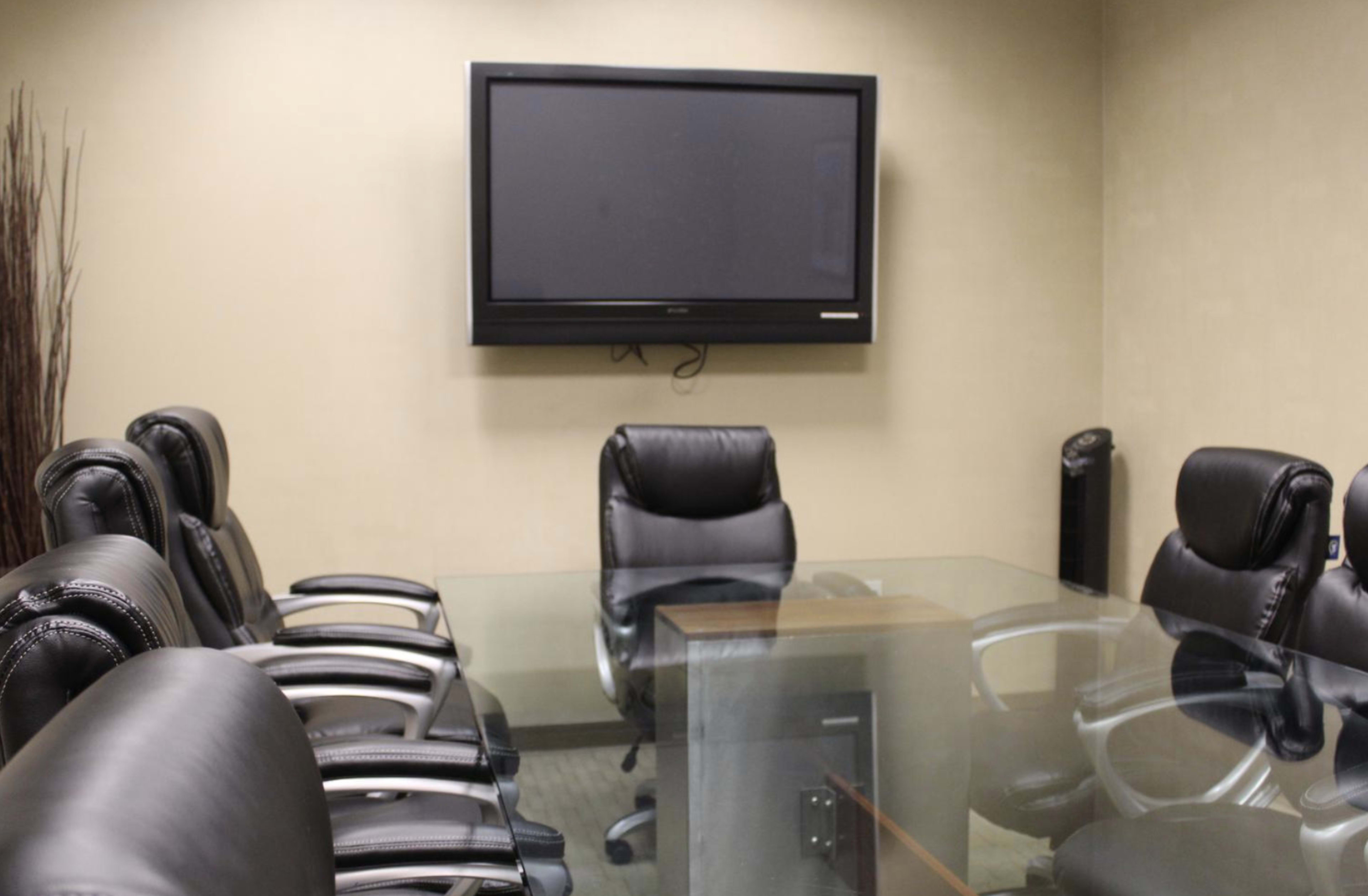 A modern conference room features a glass table surrounded by black leather chairs and a wall-mounted television.