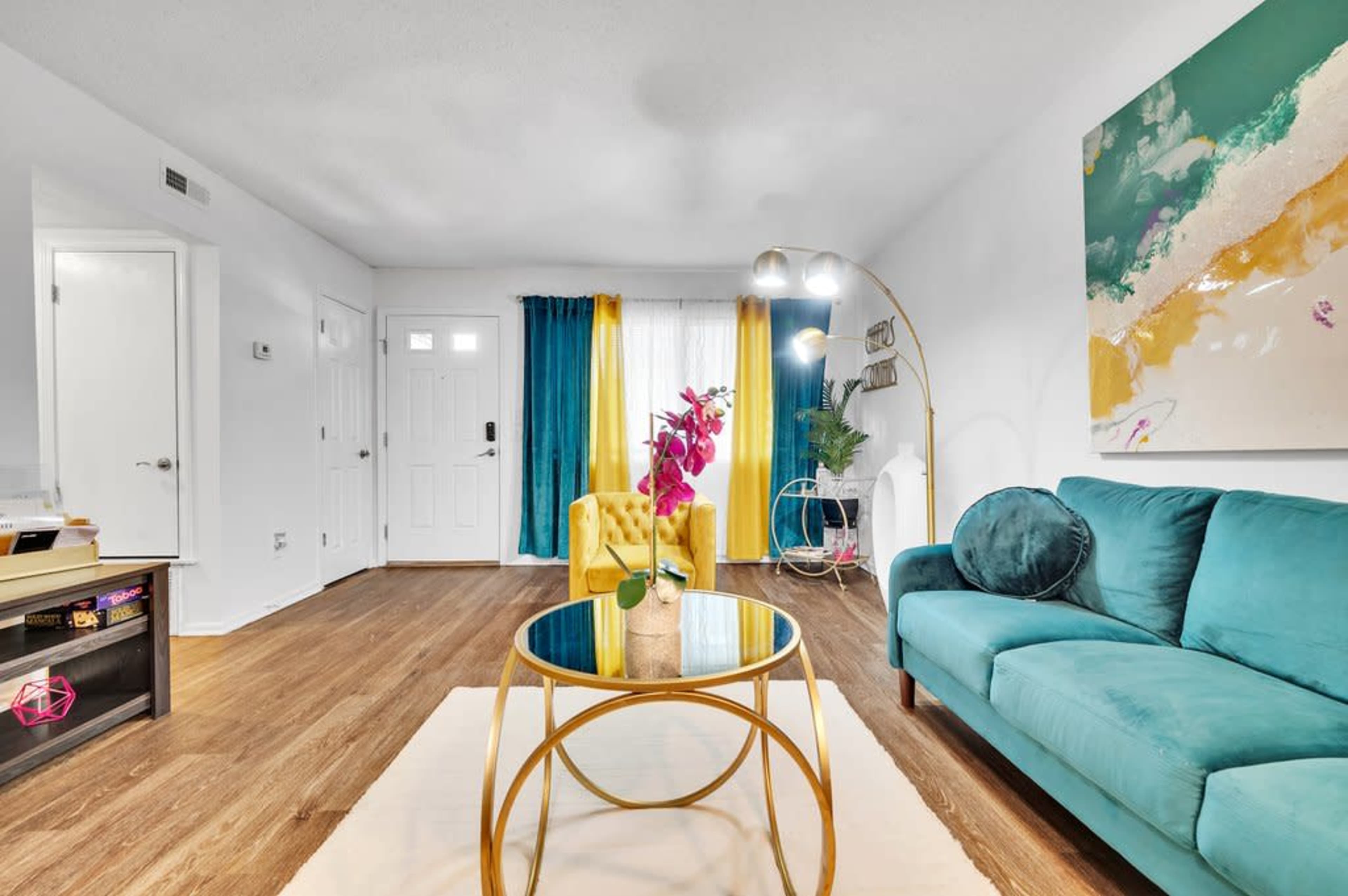 A living room features a teal couch, a yellow armchair, a round glass coffee table, and colorful curtains, with a door and a light fixture visible in the background.